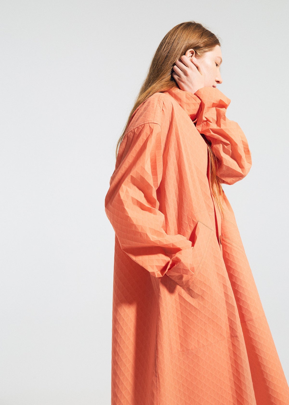 A person with long, light brown hair wears the TYPE-T 001-1 coat by A-POC ABLE ISSEY MIYAKE in coral-orange, standing sideways with one hand at their neck and the other in a pocket against a plain light background. view 8 of 12