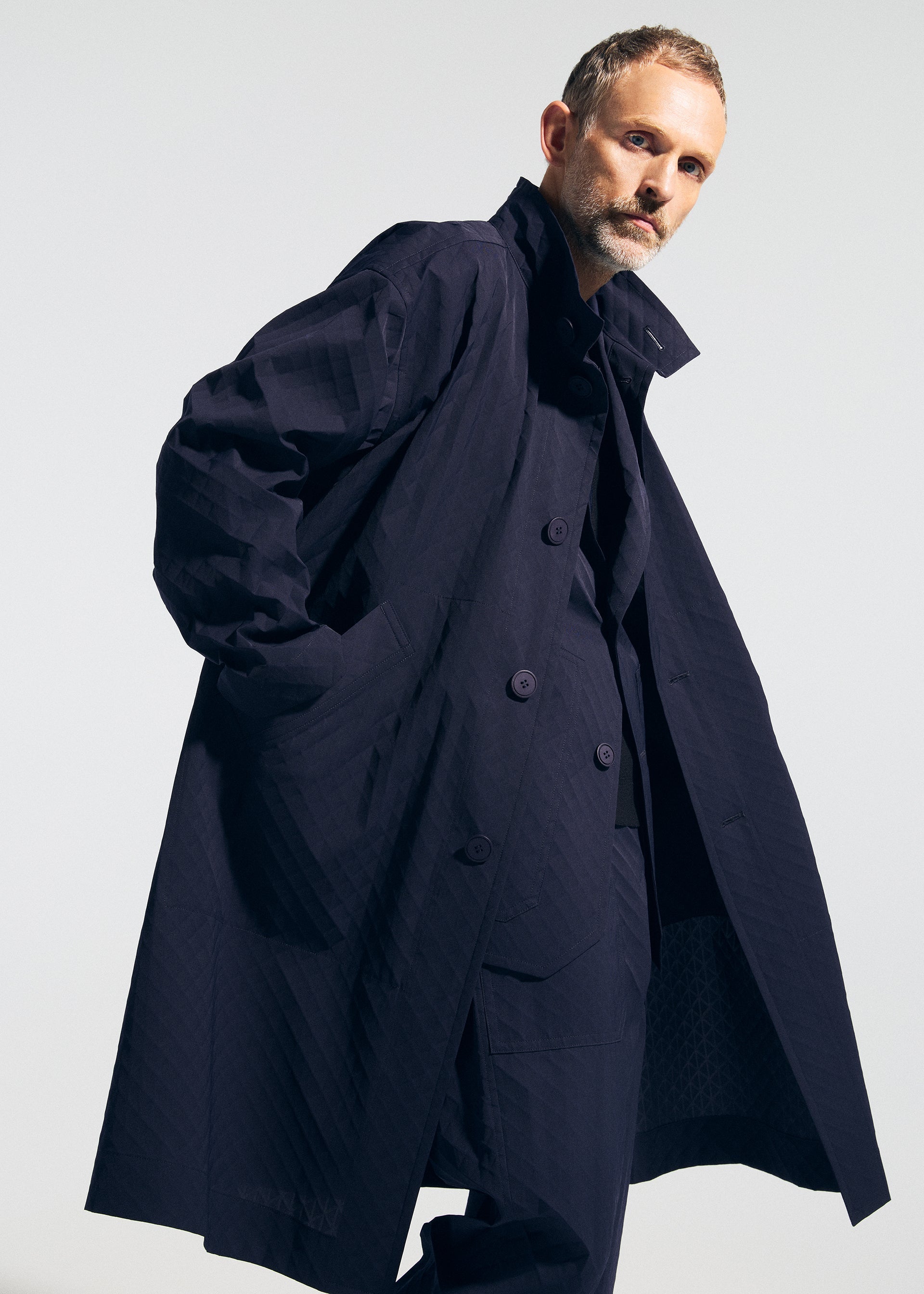 Zoomed view A man with short hair and a beard poses with his hands in the pockets, wearing the dark oversized TYPE-T 001-1 coat by A-POC ABLE ISSEY MIYAKE against a plain light background.