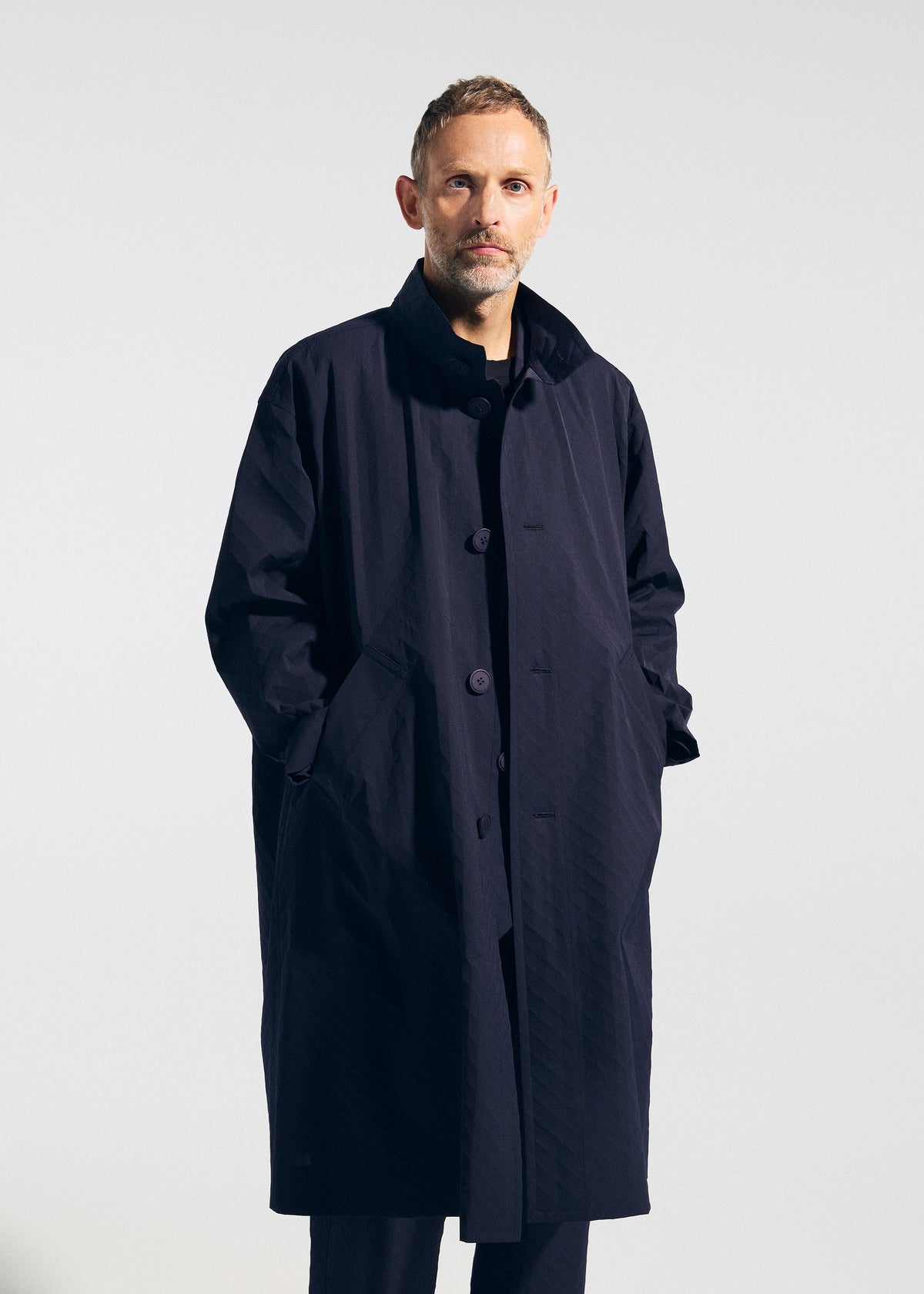 A man with short hair and a beard stands against a plain background, wearing the TYPE-T 001-1 long dark navy textured coat by A-POC ABLE ISSEY MIYAKE, with his hands in the pockets. view 10 of 12