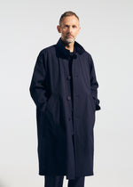 A man with short hair and a beard stands against a plain background, wearing the TYPE-T 001-1 long dark navy textured coat by A-POC ABLE ISSEY MIYAKE, with his hands in the pockets.