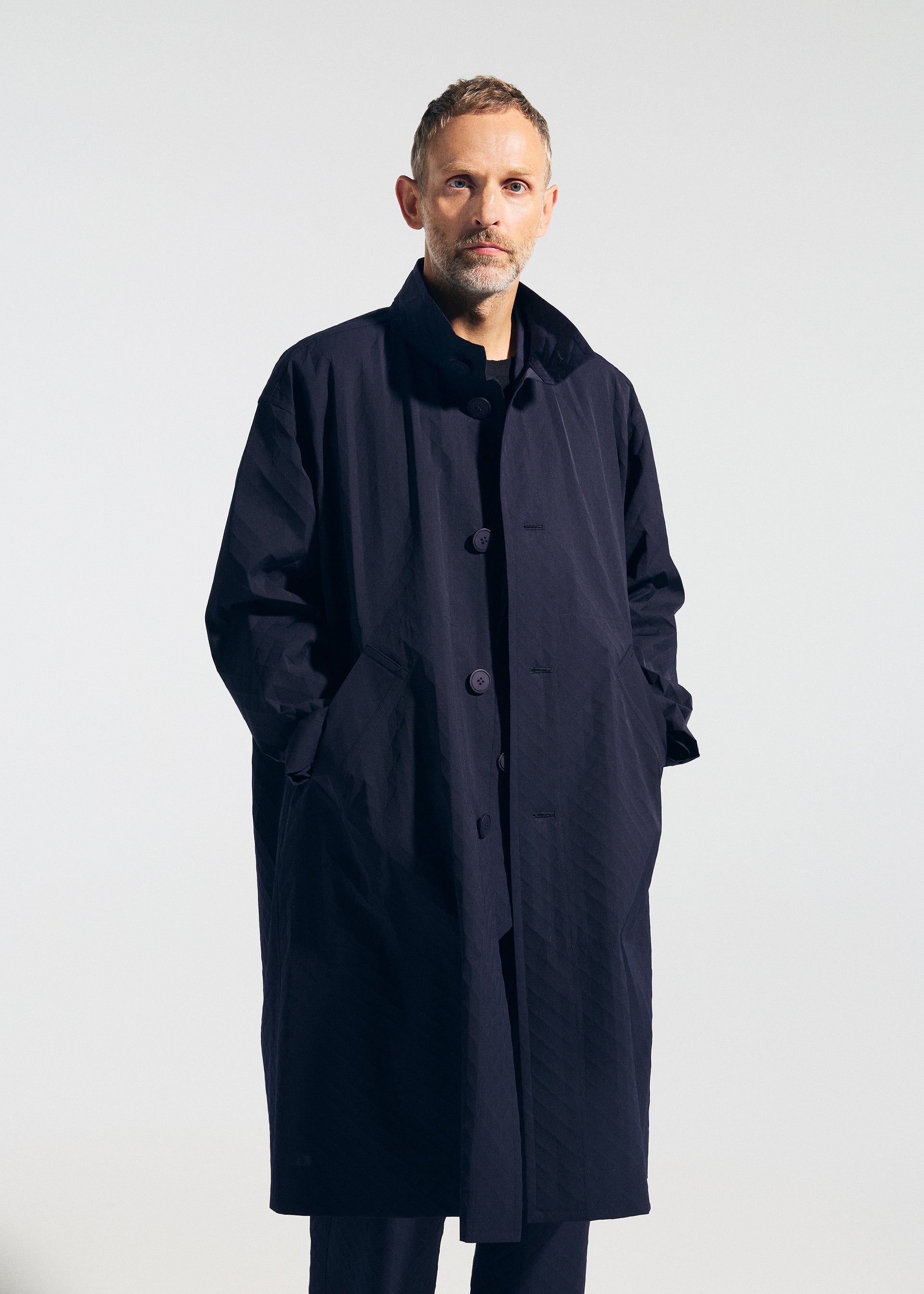 Zoomed view A man with short hair and a beard stands against a plain background, wearing the TYPE-T 001-1 long dark navy textured coat by A-POC ABLE ISSEY MIYAKE, with his hands in the pockets.