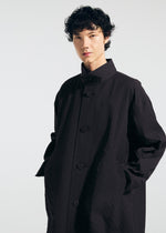 Wearing the TYPE-T 001-1 coat by A-POC ABLE ISSEY MIYAKE, a person with short, curly dark hair stands against a plain light background, one hand in their pocket and looking confidently at the camera.