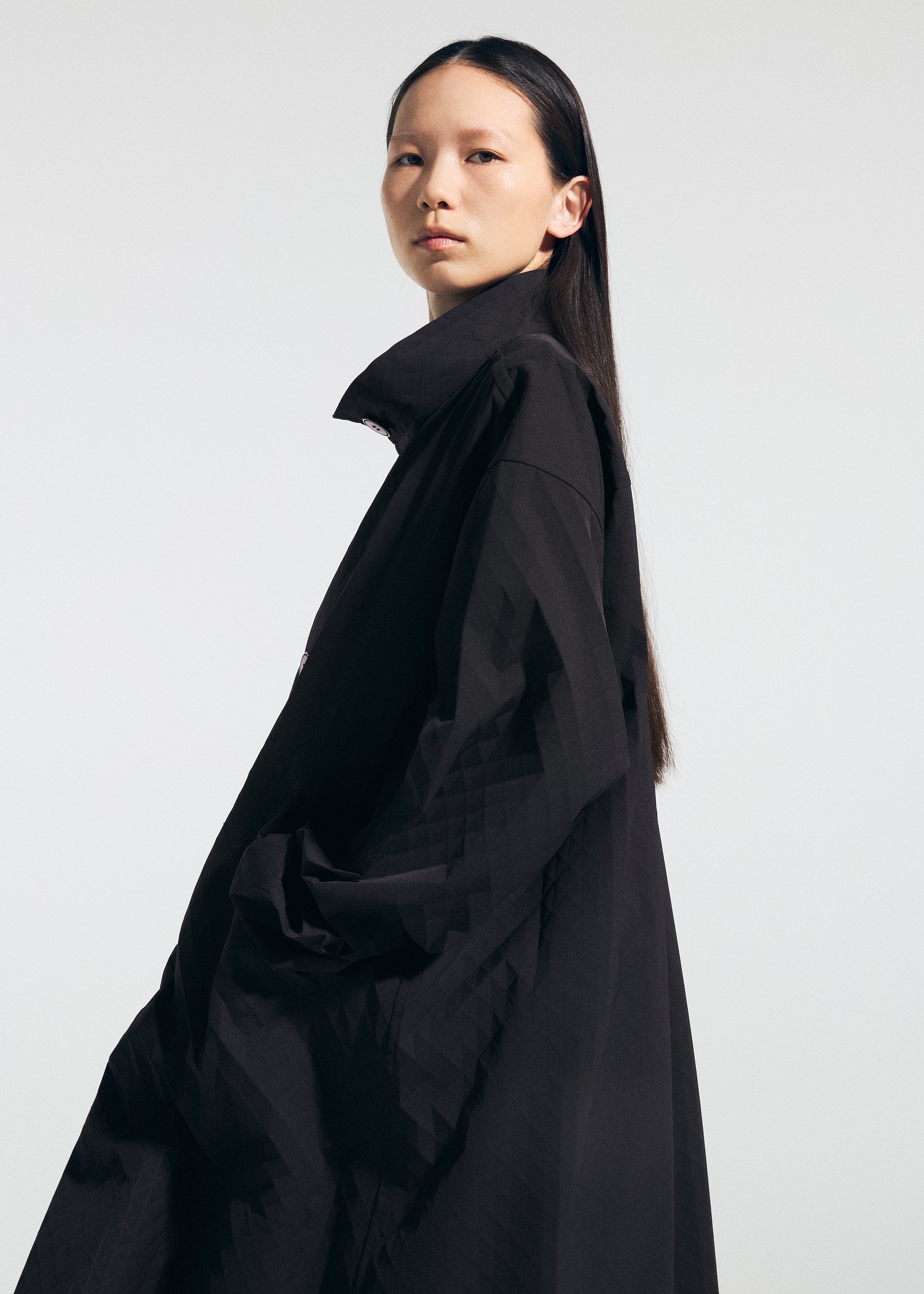 Zoomed view Wearing the TYPE-T 001-1 by A-POC ABLE ISSEY MIYAKE, a person with long, straight black hair stands against a plain light background, confidently facing the camera in a loose-fitting, subtly textured coat.