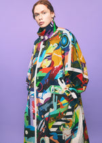 A person with red hair wears a long, colorful, abstract-patterned TYPE-XII Tomokazu Matsuyama project 003 coat by A-POC ABLE ISSEY MIYAKE, standing against a light purple background and embodying contemporary art fashion while looking confidently at the camera with hands in pockets.