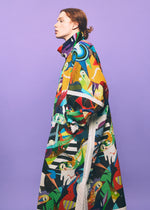 Wearing the TYPE-XII Tomokazu Matsuyama project 003 coat by A-POC ABLE ISSEY MIYAKE, a red-haired individual stands in profile against a light purple background, showcasing the garment’s long, colorful pattern inspired by contemporary art fashion.