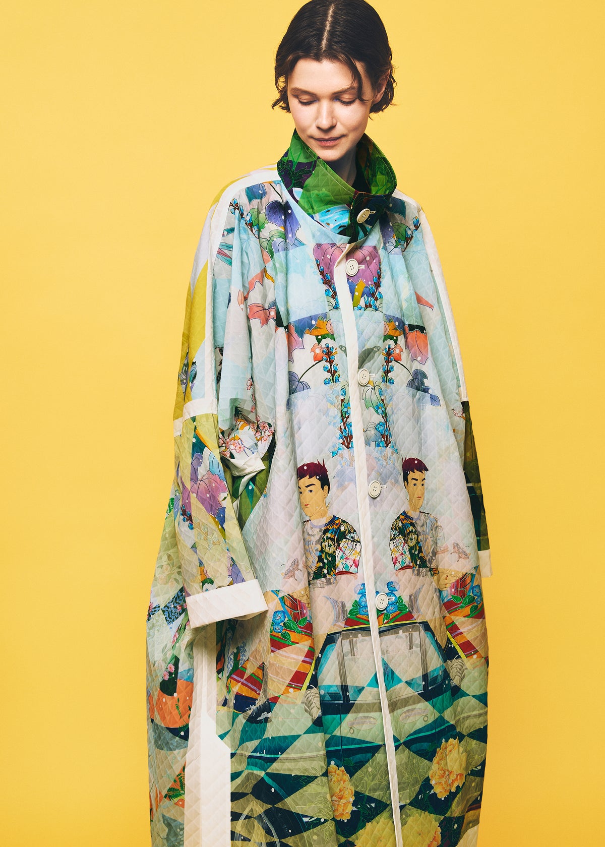 A person stands against a yellow background, wearing the TYPE-XII Tomokazu Matsuyama project 003 coat from A-POC ABLE ISSEY MIYAKE. The loose, colorful coat displays artistic patterns, geometric shapes, and illustrated faces that capture the energy of contemporary art fashion. The person looks down slightly and smiles softly. view 18 of 23