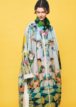 A person stands against a yellow background, wearing the TYPE-XII Tomokazu Matsuyama project 003 coat from A-POC ABLE ISSEY MIYAKE. The loose, colorful coat displays artistic patterns, geometric shapes, and illustrated faces that capture the energy of contemporary art fashion. The person looks down slightly and smiles softly.