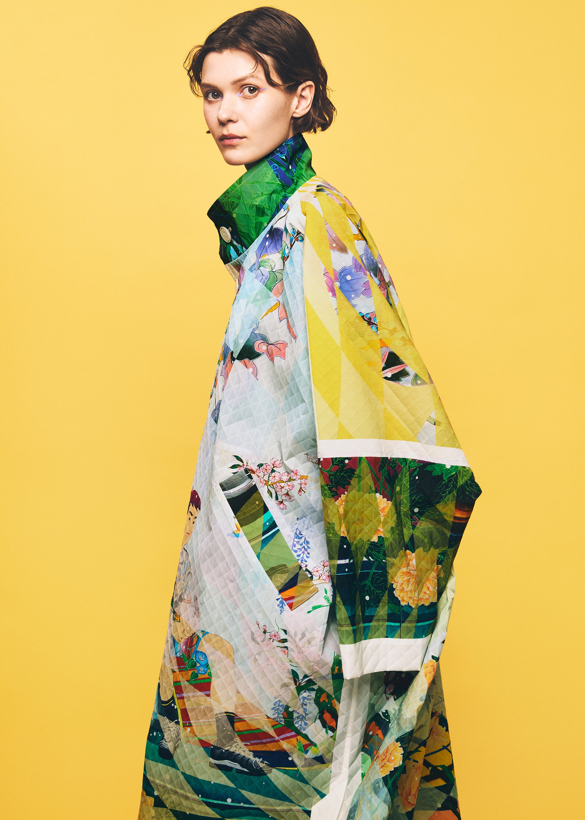 Zoomed view A person with short brown hair wears the TYPE-XII Tomokazu Matsuyama project 003 quilted coat by A-POC ABLE ISSEY MIYAKE, featuring a colorful, geometric pattern inspired by contemporary art fashion, and stands against a solid yellow background, gazing at the camera.