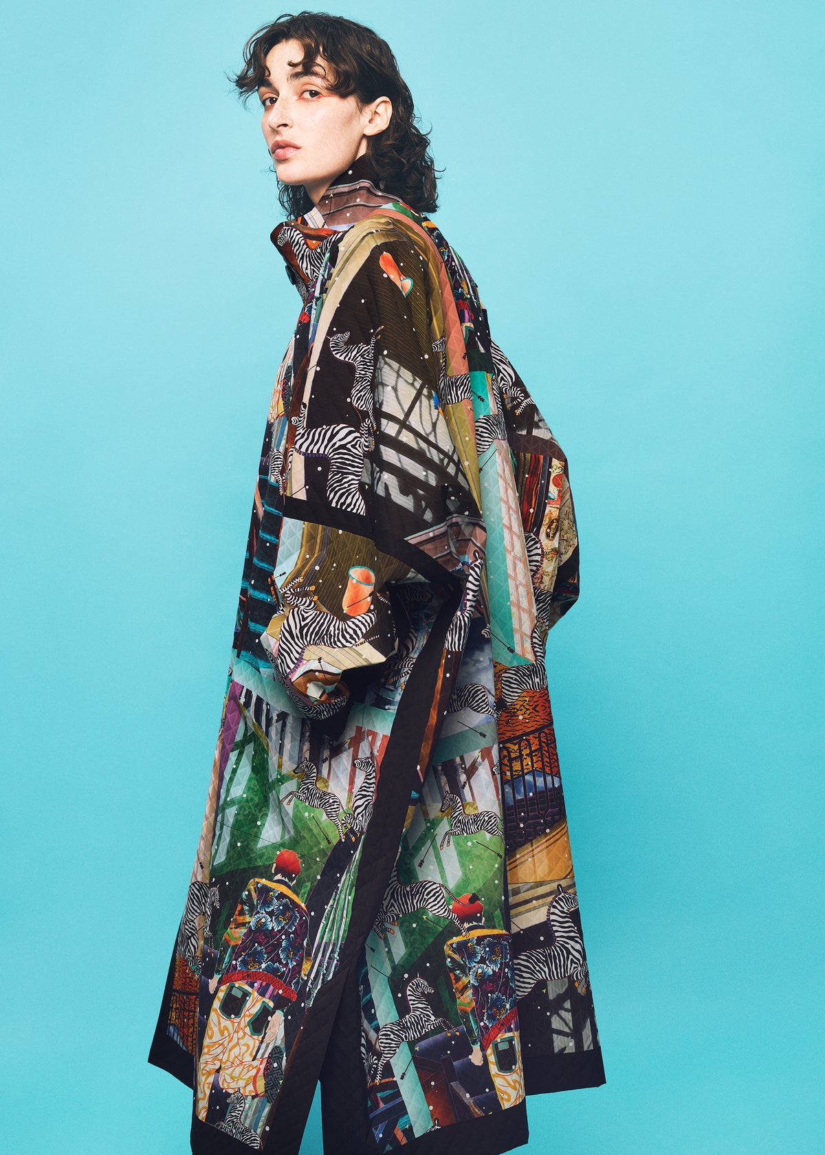 A person with curly hair wears a long, colorful patterned coat from the TYPE-XII Tomokazu Matsuyama project 003 by A-POC ABLE ISSEY MIYAKE, featuring various artistic designs, standing against a solid light blue background—a striking example of contemporary art fashion. view 21 of 23
