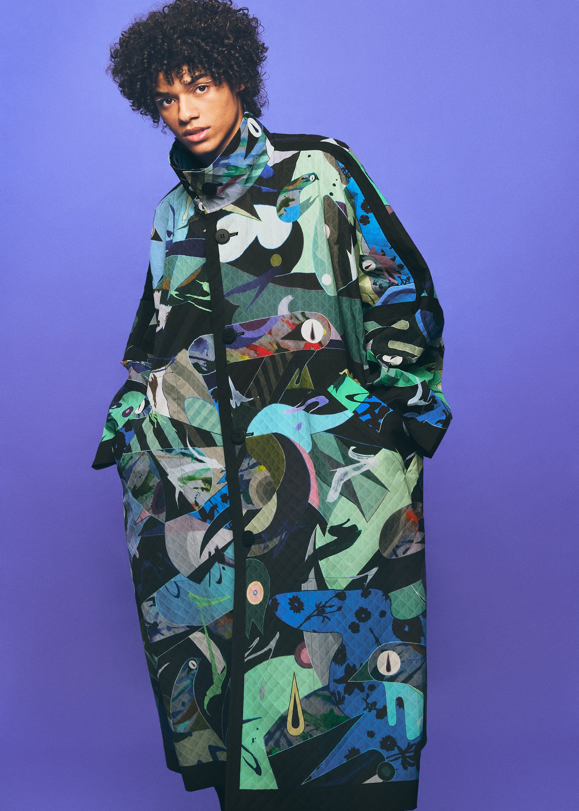 Zoomed view A person wearing a long, colorful TYPE-XII Tomokazu Matsuyama project 003 coat by A-POC ABLE ISSEY MIYAKE, featuring bold abstract patterns in green, blue, and black, stands against a purple background with hands in pockets, gazing at the camera—a striking statement of contemporary art fashion.