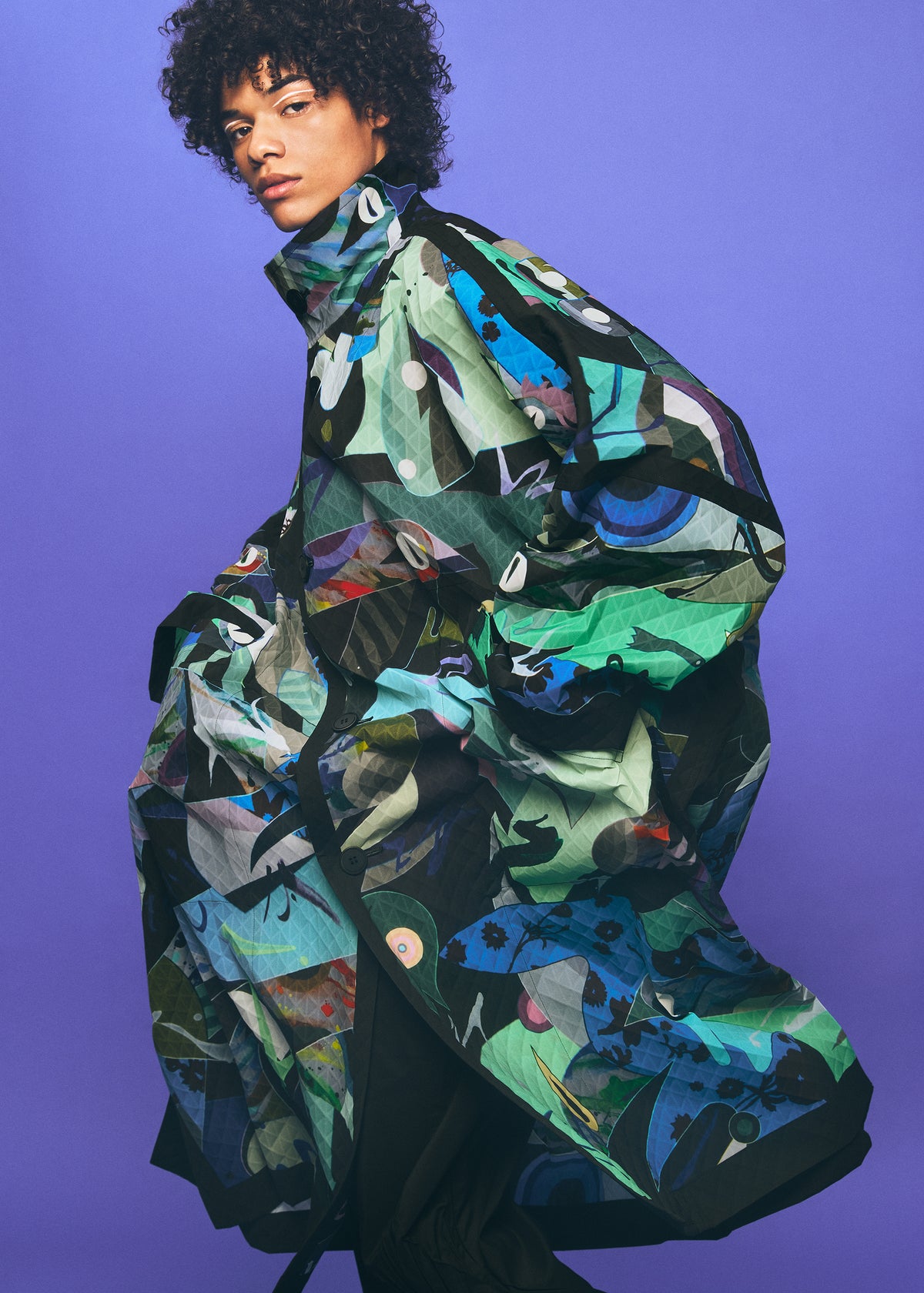 An individual with curly hair wears a vibrant, abstract-patterned oversized TYPE-XII Tomokazu Matsuyama project 003 coat from A-POC ABLE ISSEY MIYAKE, inspired by contemporary art fashion, standing confidently against a solid purple background and gazing directly at the camera. view 23 of 23