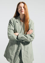 A person with long red hair stands facing the camera, arms crossed, wearing the TYPE-U 001 light green oversized, wrinkle-resistant blazer and matching pants by A-POC ABLE ISSEY MIYAKE over a white shirt, against a plain light background.