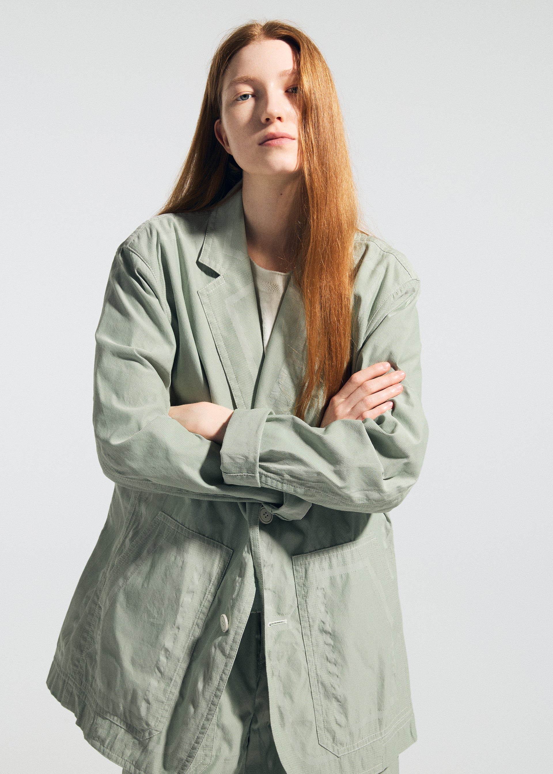 Zoomed view A person with long red hair stands facing the camera, arms crossed, wearing the TYPE-U 001 light green oversized, wrinkle-resistant blazer and matching pants by A-POC ABLE ISSEY MIYAKE over a white shirt, against a plain light background.
