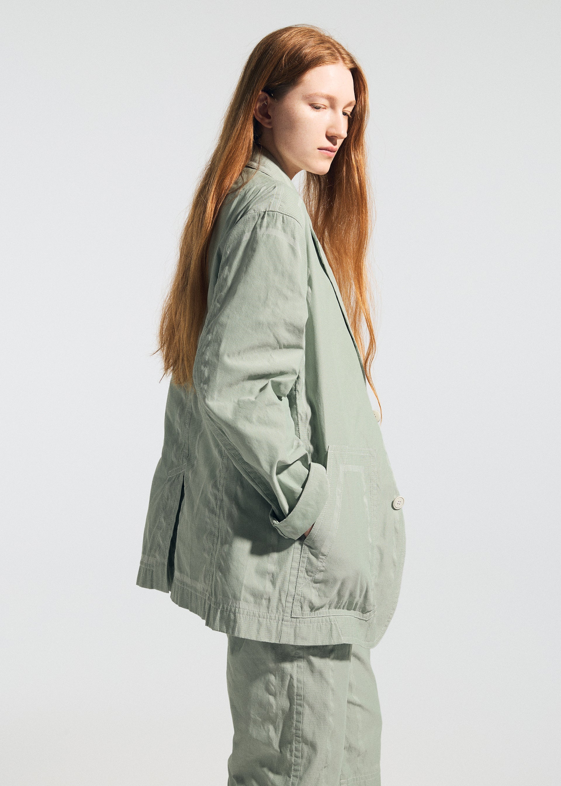 Zoomed view A person with long red hair stands in profile against a plain white background, exuding relaxed comfort in the light green, oversized TYPE-U 001 jacket and matching pants from A-POC ABLE ISSEY MIYAKE, hands tucked casually into the jacket’s pockets.