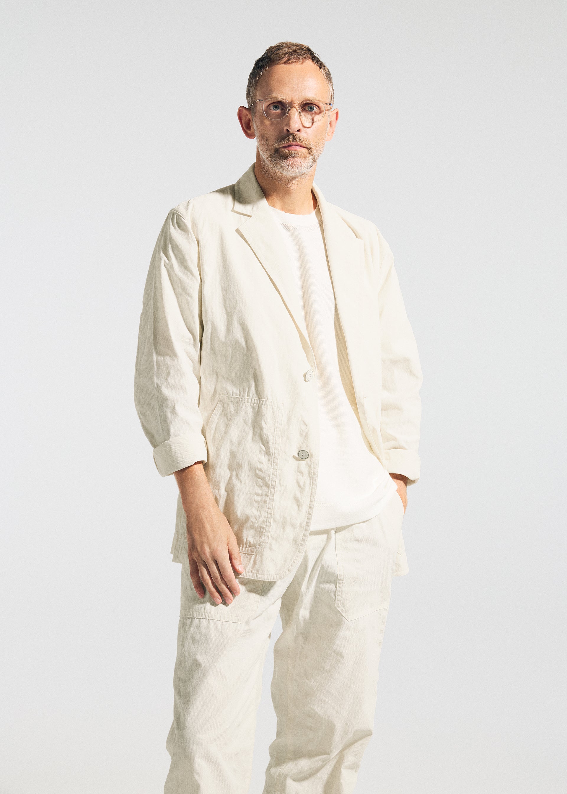 Zoomed view A person with short hair, glasses, and a beard stands against a plain background, wearing the TYPE-U 001 jacket by A-POC ABLE ISSEY MIYAKE—a light-colored, wrinkle-resistant piece that drapes with relaxed comfort over their shirt and pants.
