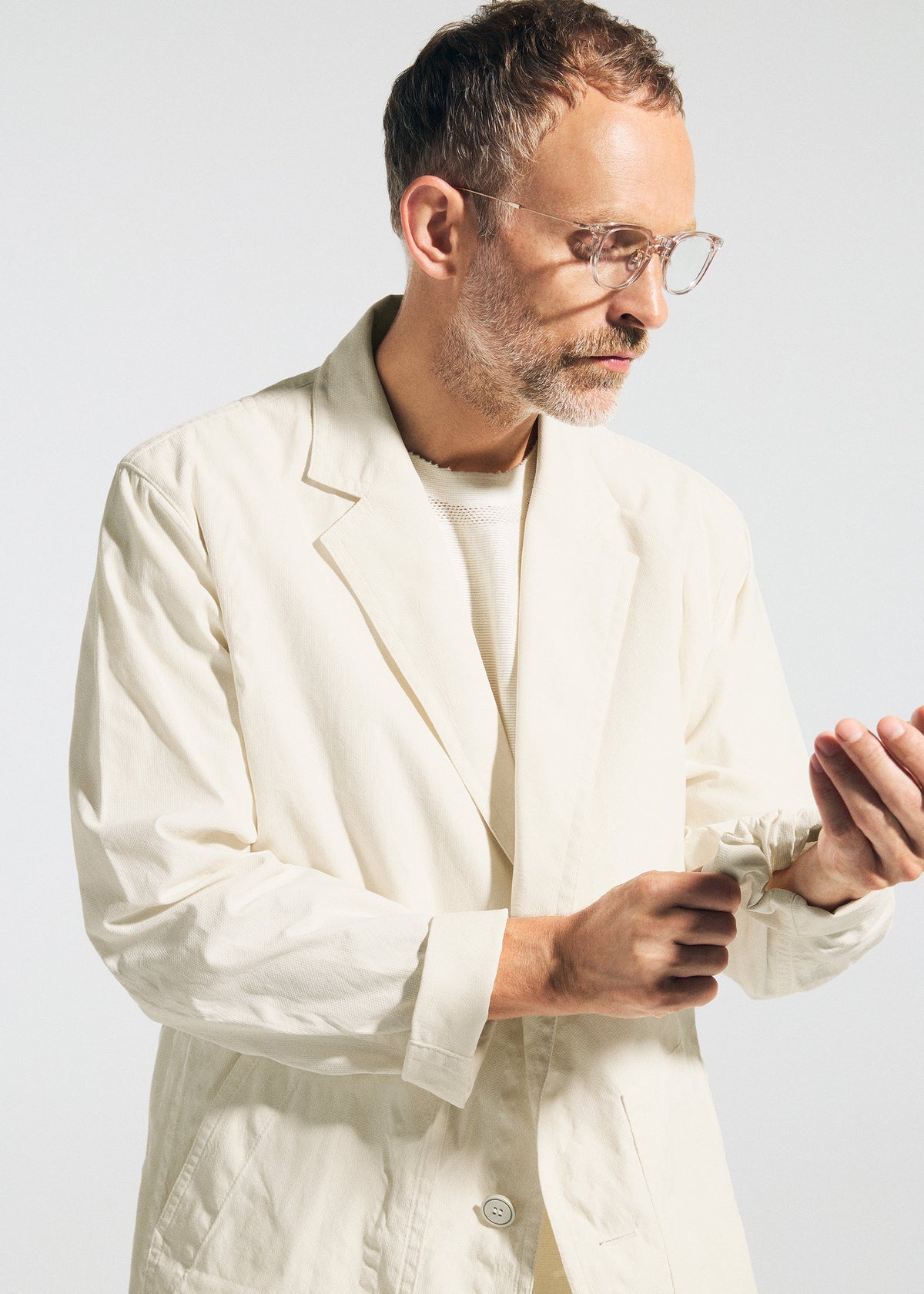 A man with short hair, a beard, and glasses wears the cream-colored TYPE-U 001 wrinkle-resistant blazer from A-POC ABLE ISSEY MIYAKE, looking down as he adjusts his sleeve against a plain light background. view 9 of 11