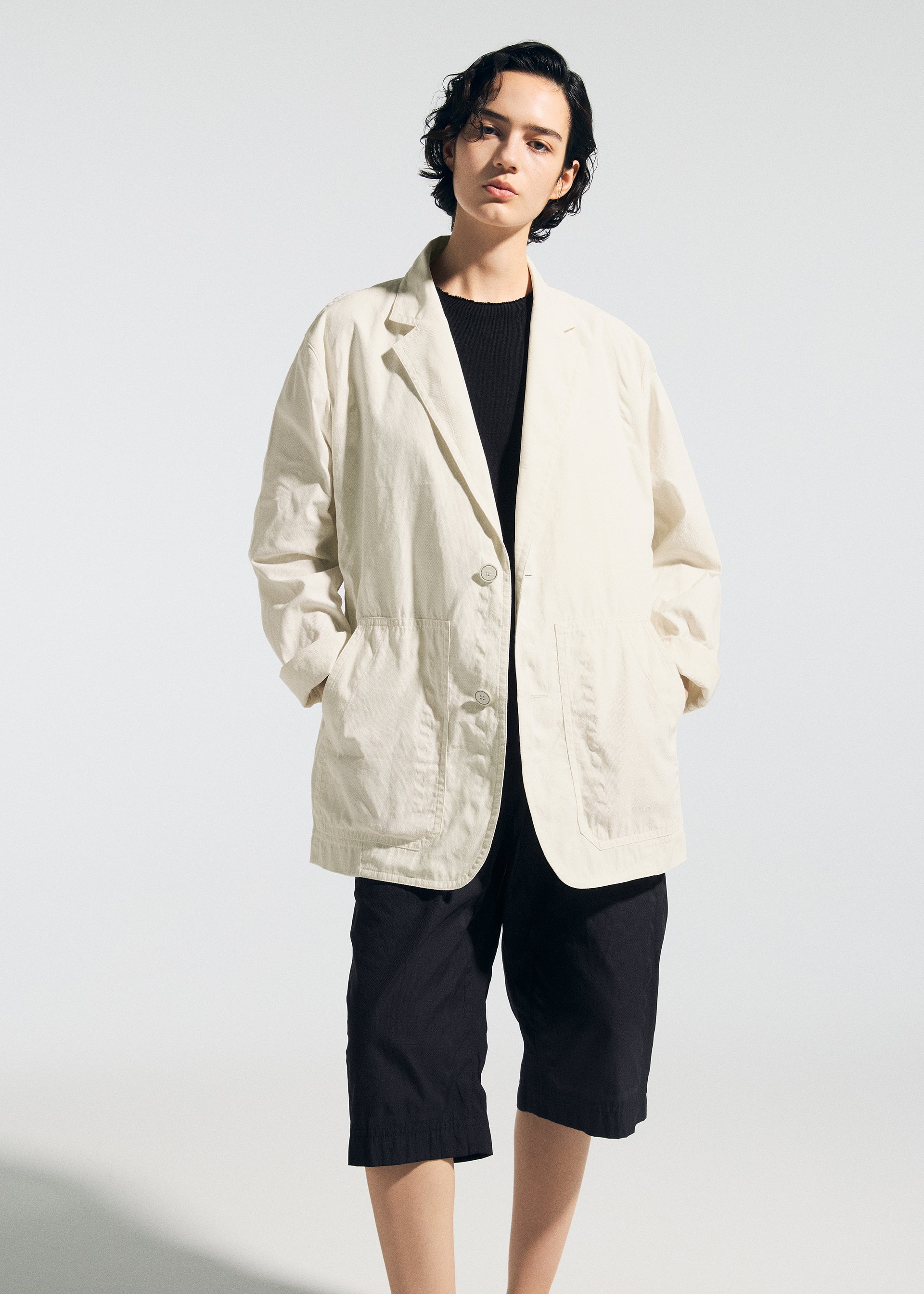 Zoomed view A person with short dark hair wears the TYPE-U 001 by A-POC ABLE ISSEY MIYAKE—a loose, cream-colored shape-memory jacket layered over a black shirt and black knee-length shorts—standing against a plain, light backdrop and exemplifying relaxed comfort with the effortless, wrinkle-resistant style of the collection.