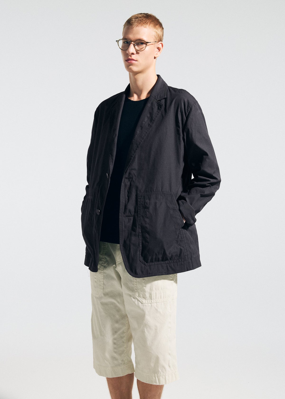 A person wearing glasses and the TYPE-U 001 jacket from A-POC ABLE ISSEY MIYAKE—a black, wrinkle-resistant piece with shape-memory properties—over a dark shirt and paired with light-colored knee-length shorts stands against a plain, light background, embodying relaxed comfort. view 11 of 11