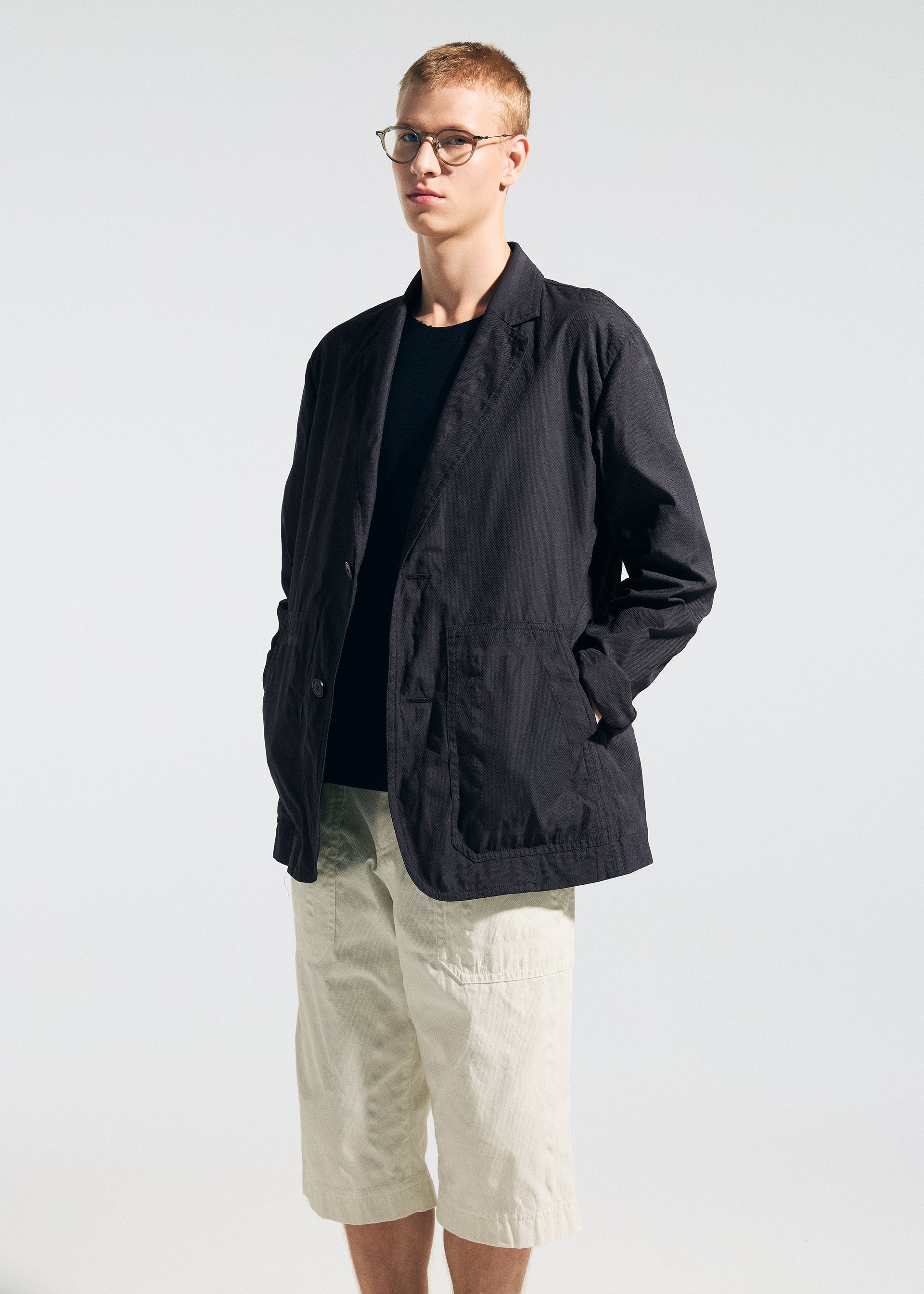 Zoomed view A person wearing glasses and the TYPE-U 001 jacket from A-POC ABLE ISSEY MIYAKE—a black, wrinkle-resistant piece with shape-memory properties—over a dark shirt and paired with light-colored knee-length shorts stands against a plain, light background, embodying relaxed comfort.
