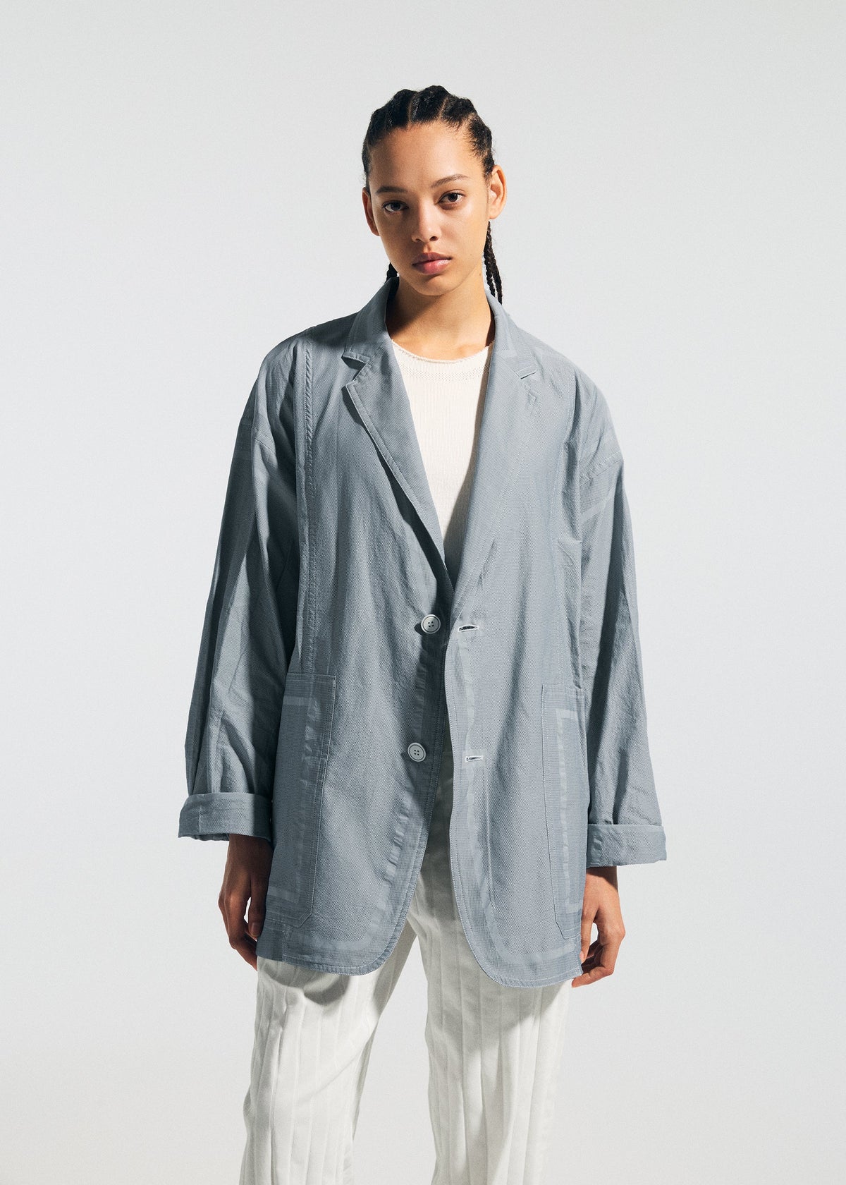 A person with braided hair models the TYPE-U 002 light gray wrinkle-resistant oversized blazer by A-POC ABLE ISSEY MIYAKE over a white shirt and pants, set against a plain light background. view 4 of 7