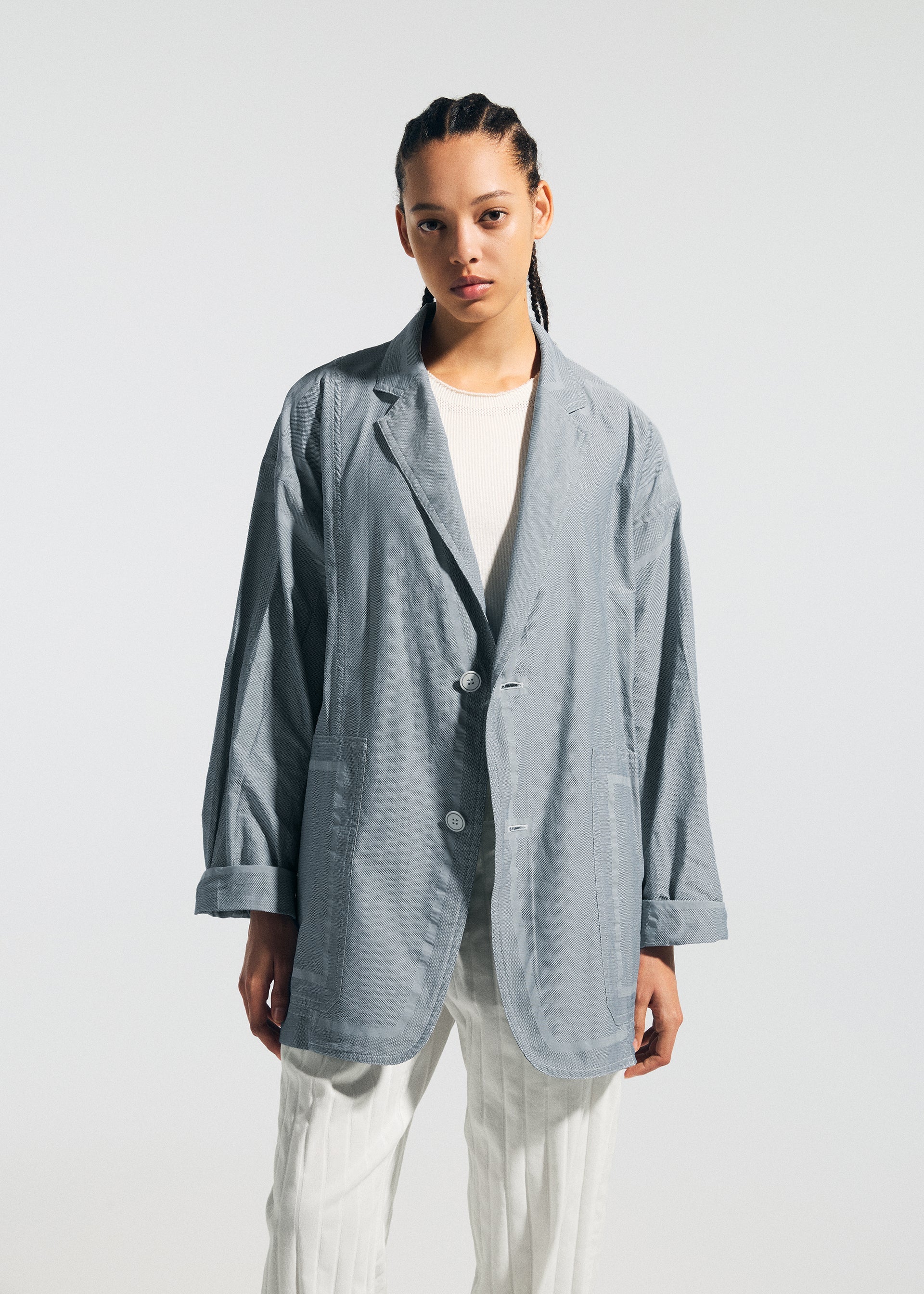 Zoomed view A person with braided hair models the TYPE-U 002 light gray wrinkle-resistant oversized blazer by A-POC ABLE ISSEY MIYAKE over a white shirt and pants, set against a plain light background.