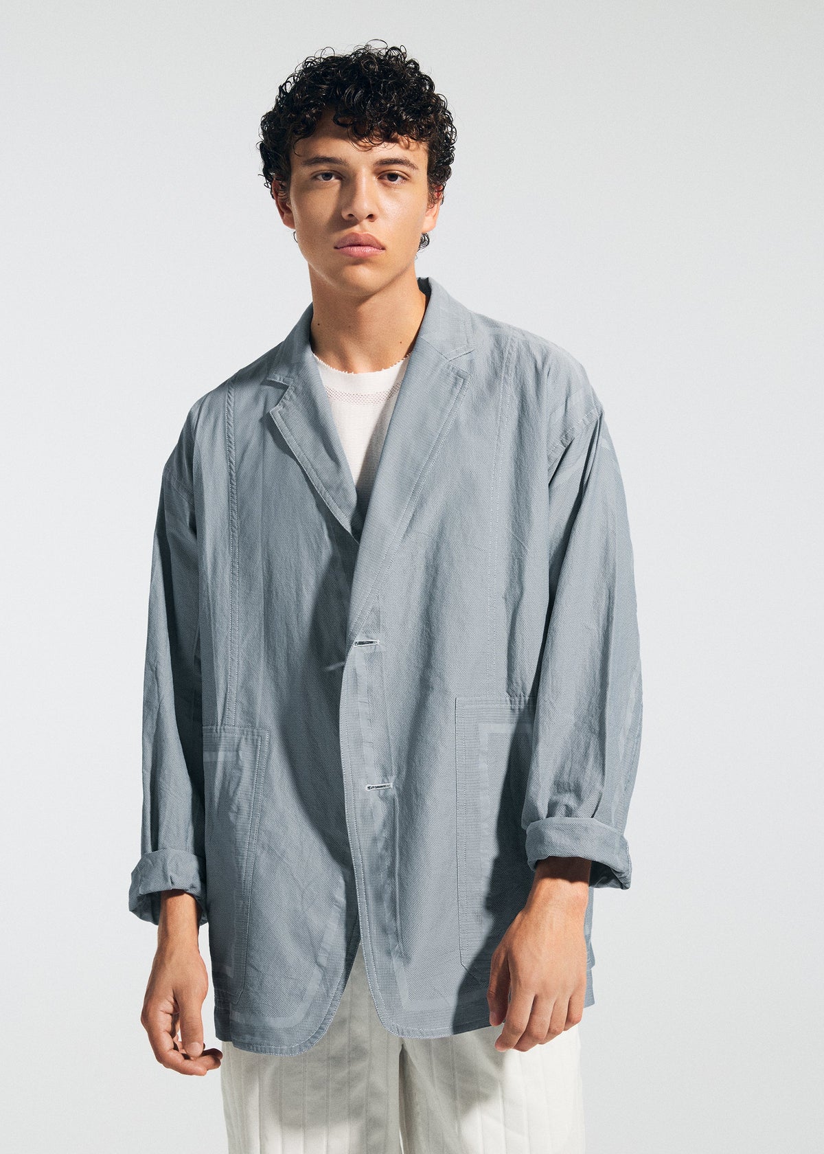 A young man with curly dark hair wears the TYPE-U 002 oversized light gray blazer by A-POC ABLE ISSEY MIYAKE over a white shirt and pants, standing against a plain light background with a neutral expression. view 5 of 7