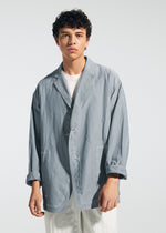 A young man with curly dark hair wears the TYPE-U 002 oversized light gray blazer by A-POC ABLE ISSEY MIYAKE over a white shirt and pants, standing against a plain light background with a neutral expression.