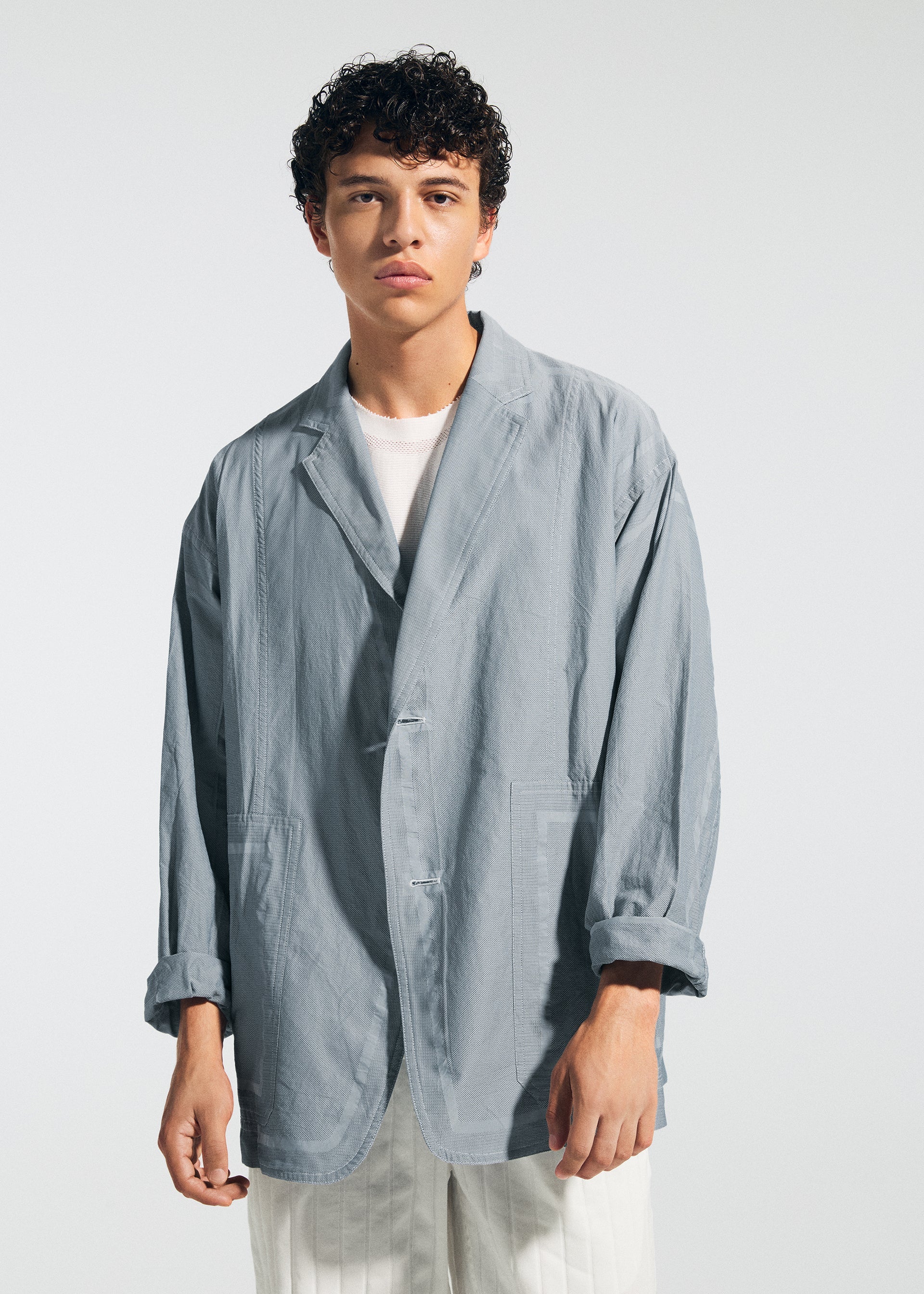 Zoomed view A young man with curly dark hair wears the TYPE-U 002 oversized light gray blazer by A-POC ABLE ISSEY MIYAKE over a white shirt and pants, standing against a plain light background with a neutral expression.