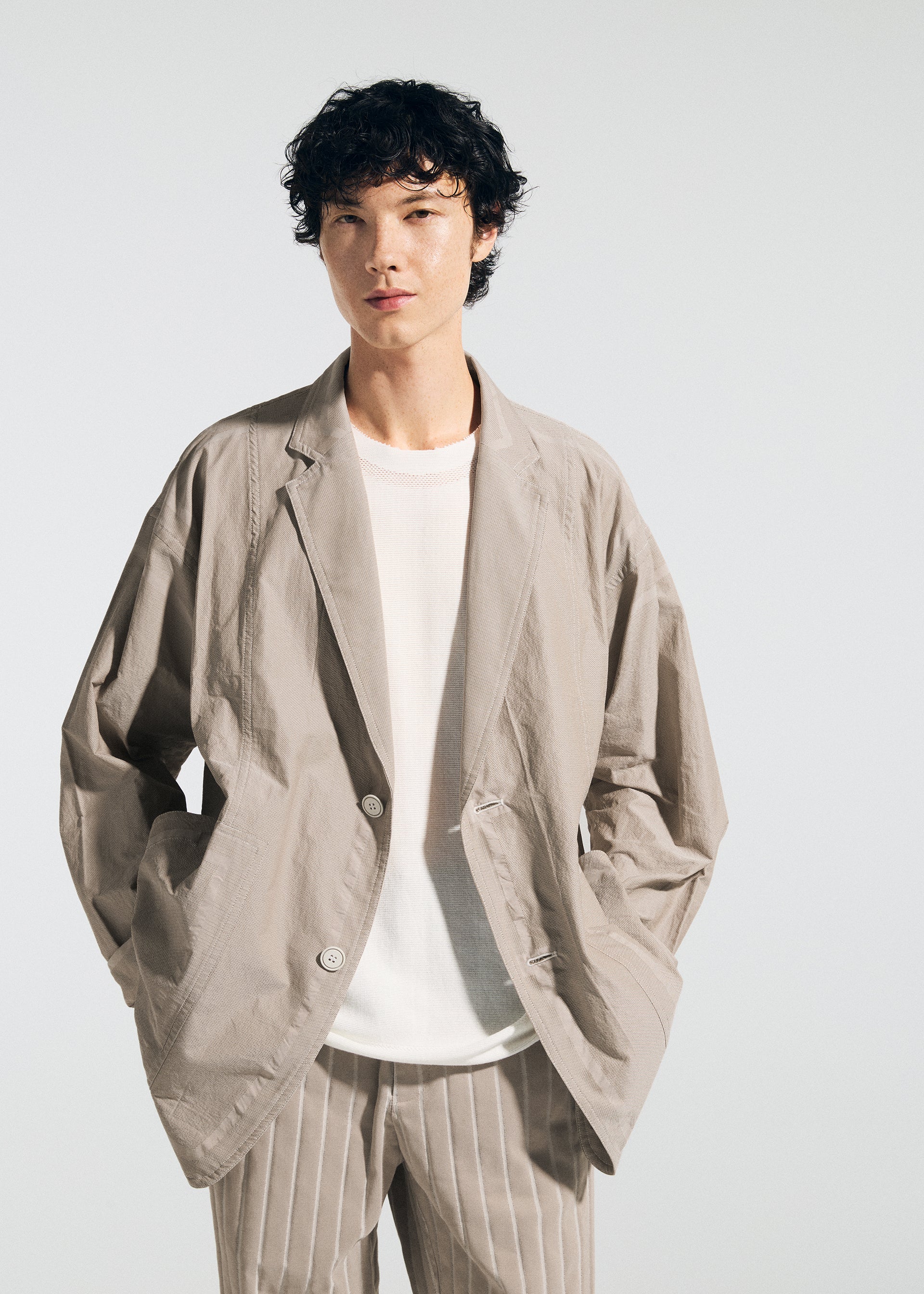 Zoomed view A person with short, curly dark hair wears the TYPE-U 002 beige blazer by A-POC ABLE ISSEY MIYAKE over a white shirt and beige pinstriped pants, standing against a plain light background with hands in blazer pockets.