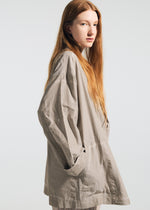 A young woman with long red hair stands sideways, hands in the pockets of the wrinkle-resistant beige TYPE-U 002 jacket by A-POC ABLE ISSEY MIYAKE, set against a plain light background.