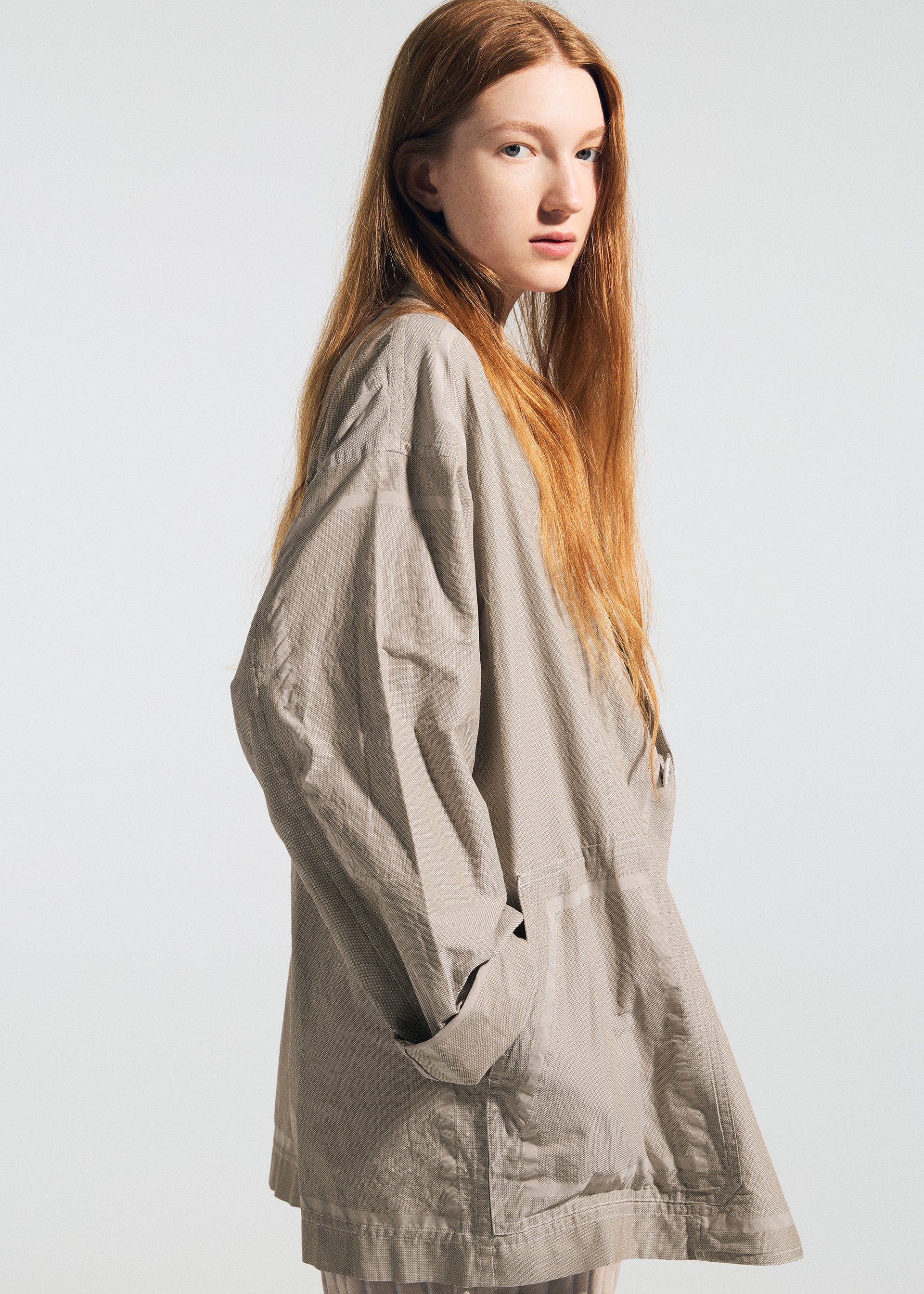Zoomed view A young woman with long red hair stands sideways, hands in the pockets of the wrinkle-resistant beige TYPE-U 002 jacket by A-POC ABLE ISSEY MIYAKE, set against a plain light background.