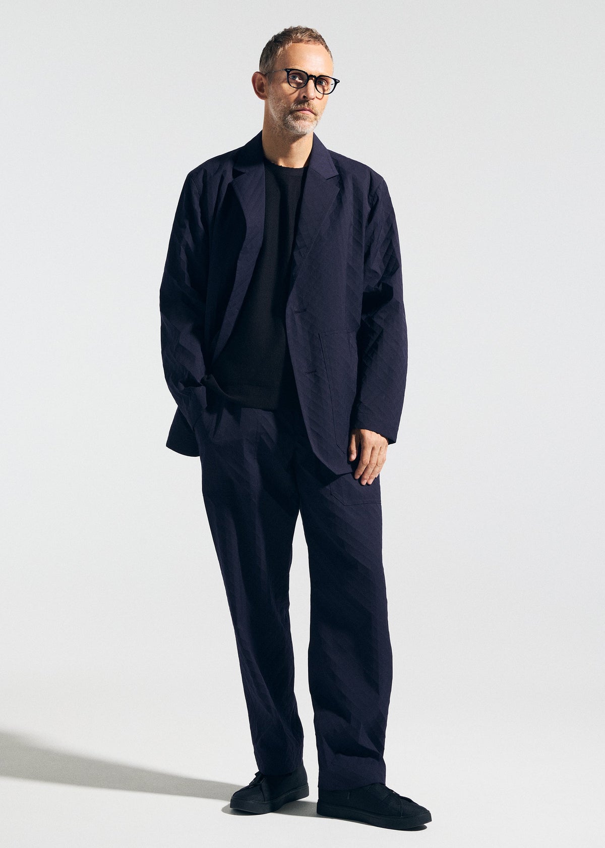 A man with short gray hair and glasses wears the A-POC ABLE ISSEY MIYAKE TYPE-T 001-2, a loose-fitting textured dark navy jacket over a black shirt, standing with one hand in his pocket against a plain light background. view 5 of 8