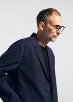 A man with short light brown hair, a beard, and glasses models the TYPE-T 001-2 dark blue textured blazer by A-POC ABLE ISSEY MIYAKE over a black shirt, standing against a plain light background looking slightly downward.