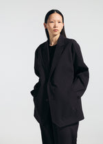 A person with straight, long dark hair faces the camera against a plain light background, wearing the oversized TYPE-T 001-2 jacket and matching pants by A-POC ABLE ISSEY MIYAKE, hands in blazer pockets and a neutral expression.