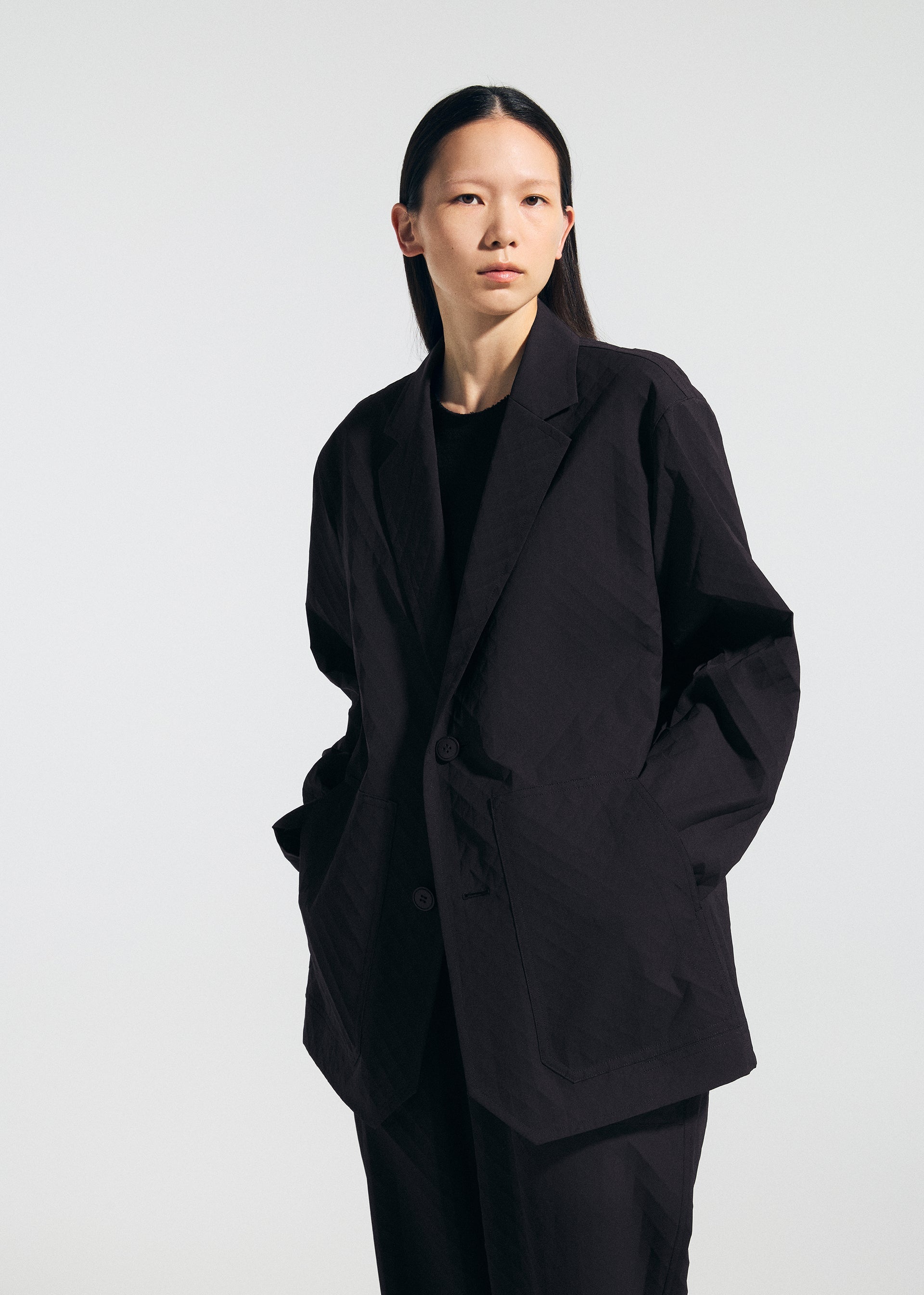 Zoomed view A person with straight, long dark hair faces the camera against a plain light background, wearing the oversized TYPE-T 001-2 jacket and matching pants by A-POC ABLE ISSEY MIYAKE, hands in blazer pockets and a neutral expression.
