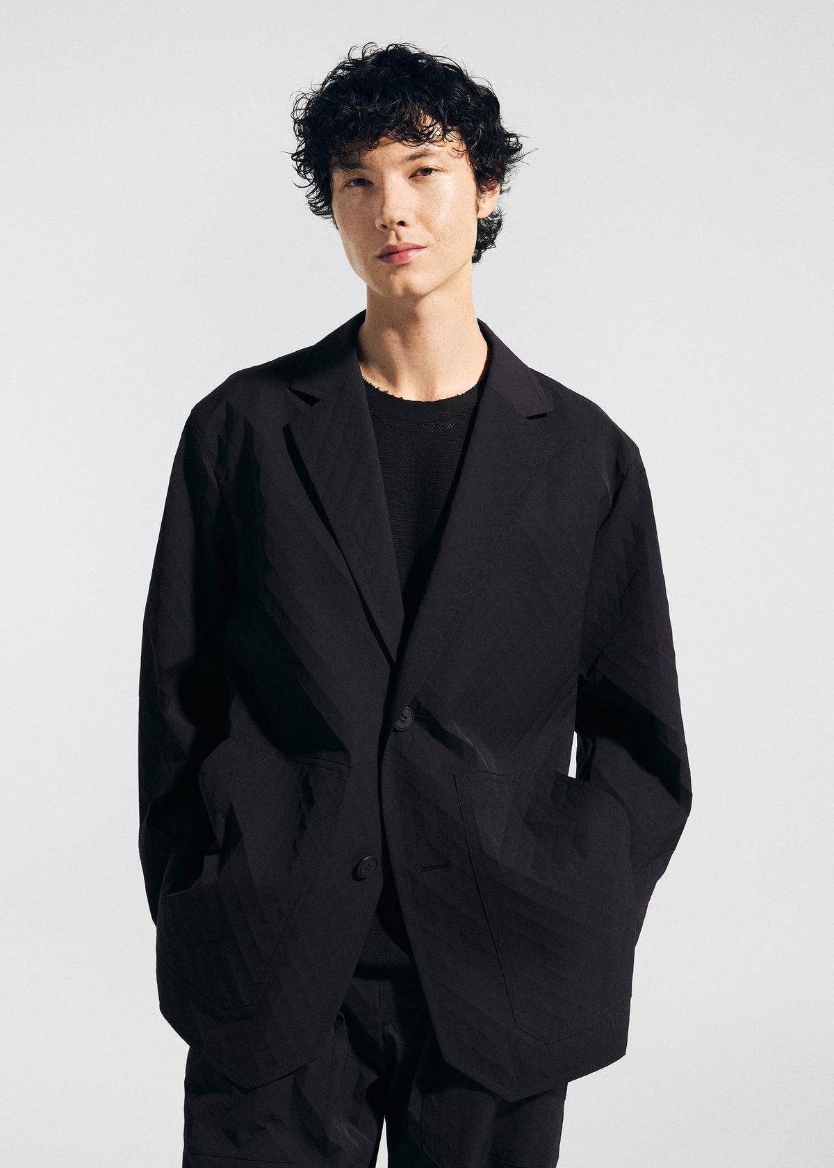 Wearing the TYPE-T 001-2 jacket by A-POC ABLE ISSEY MIYAKE over a black shirt, a person with short, curly dark hair stands against a plain light background with hands in their jacket pockets. view 8 of 8