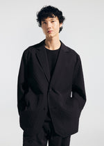 Wearing the TYPE-T 001-2 jacket by A-POC ABLE ISSEY MIYAKE over a black shirt, a person with short, curly dark hair stands against a plain light background with hands in their jacket pockets.