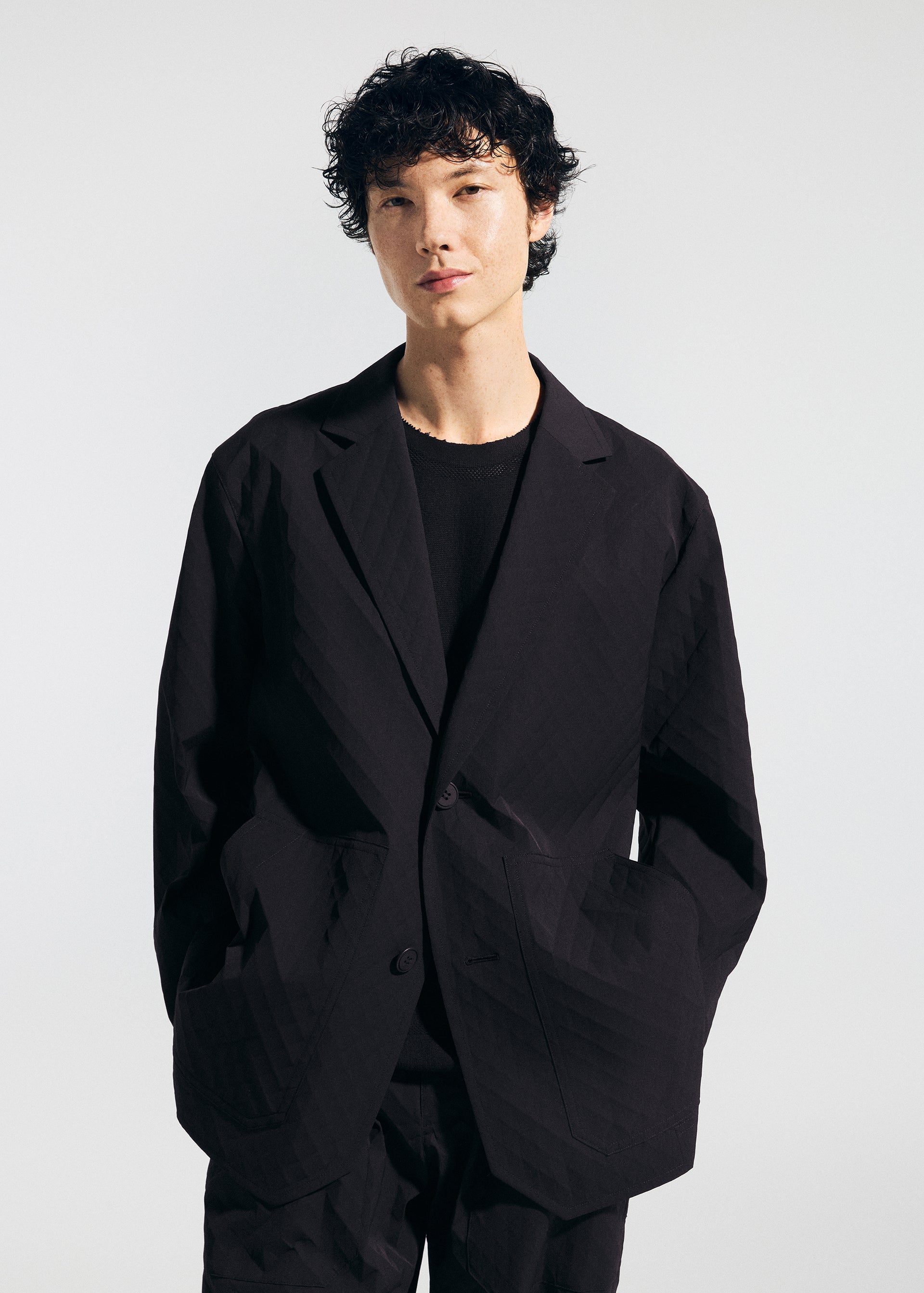 Zoomed view Wearing the TYPE-T 001-2 jacket by A-POC ABLE ISSEY MIYAKE over a black shirt, a person with short, curly dark hair stands against a plain light background with hands in their jacket pockets.