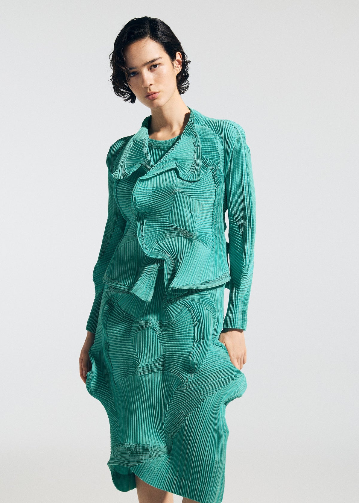 A person with short dark hair models the TYPE-O 003 teal dress by A-POC ABLE ISSEY MIYAKE, made from recycled polyester with layered, pleated fabric for a 3D wavy effect, standing against a plain white background. view 6 of 7