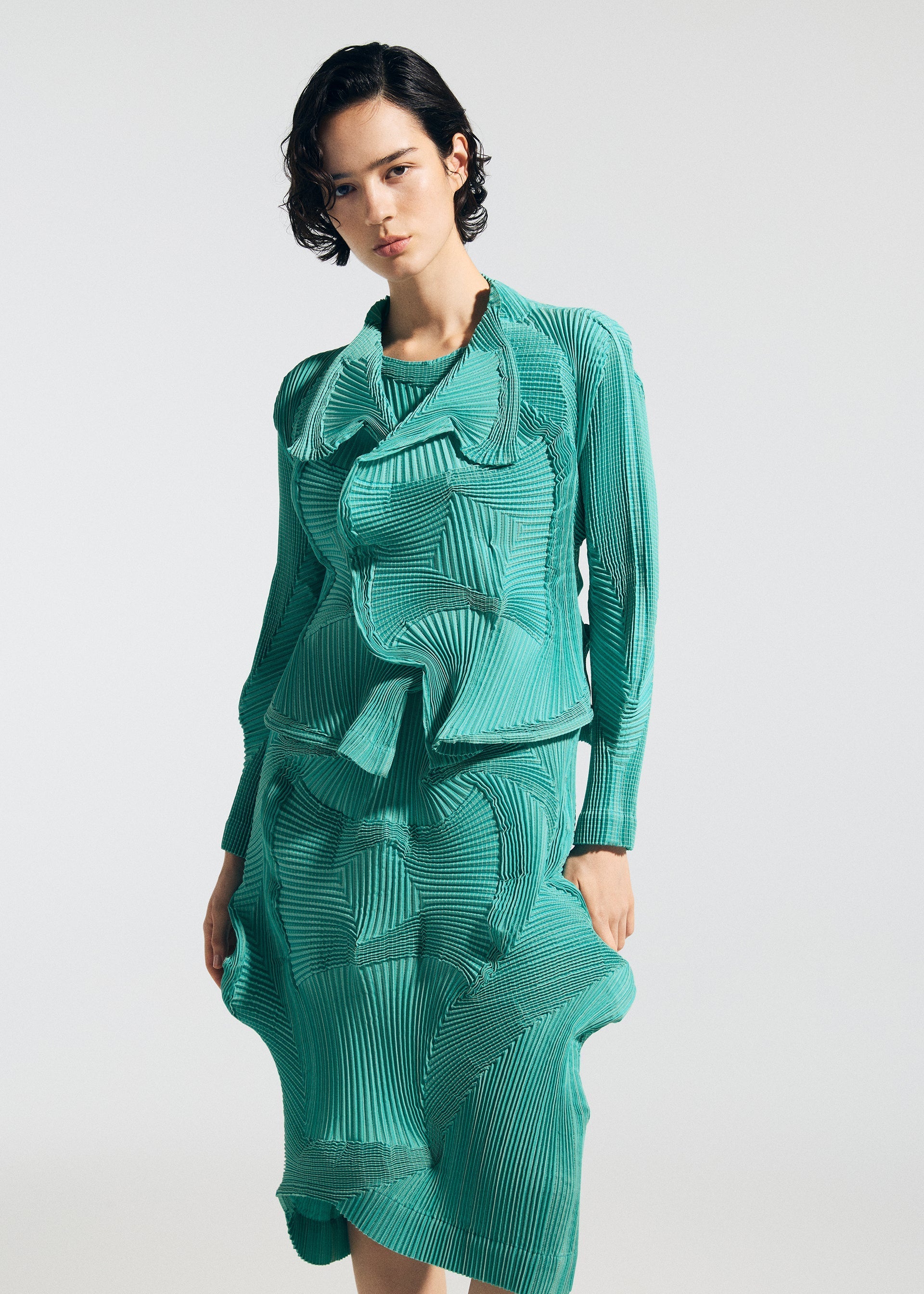 Zoomed view A person with short dark hair models the TYPE-O 003 teal dress by A-POC ABLE ISSEY MIYAKE, made from recycled polyester with layered, pleated fabric for a 3D wavy effect, standing against a plain white background.