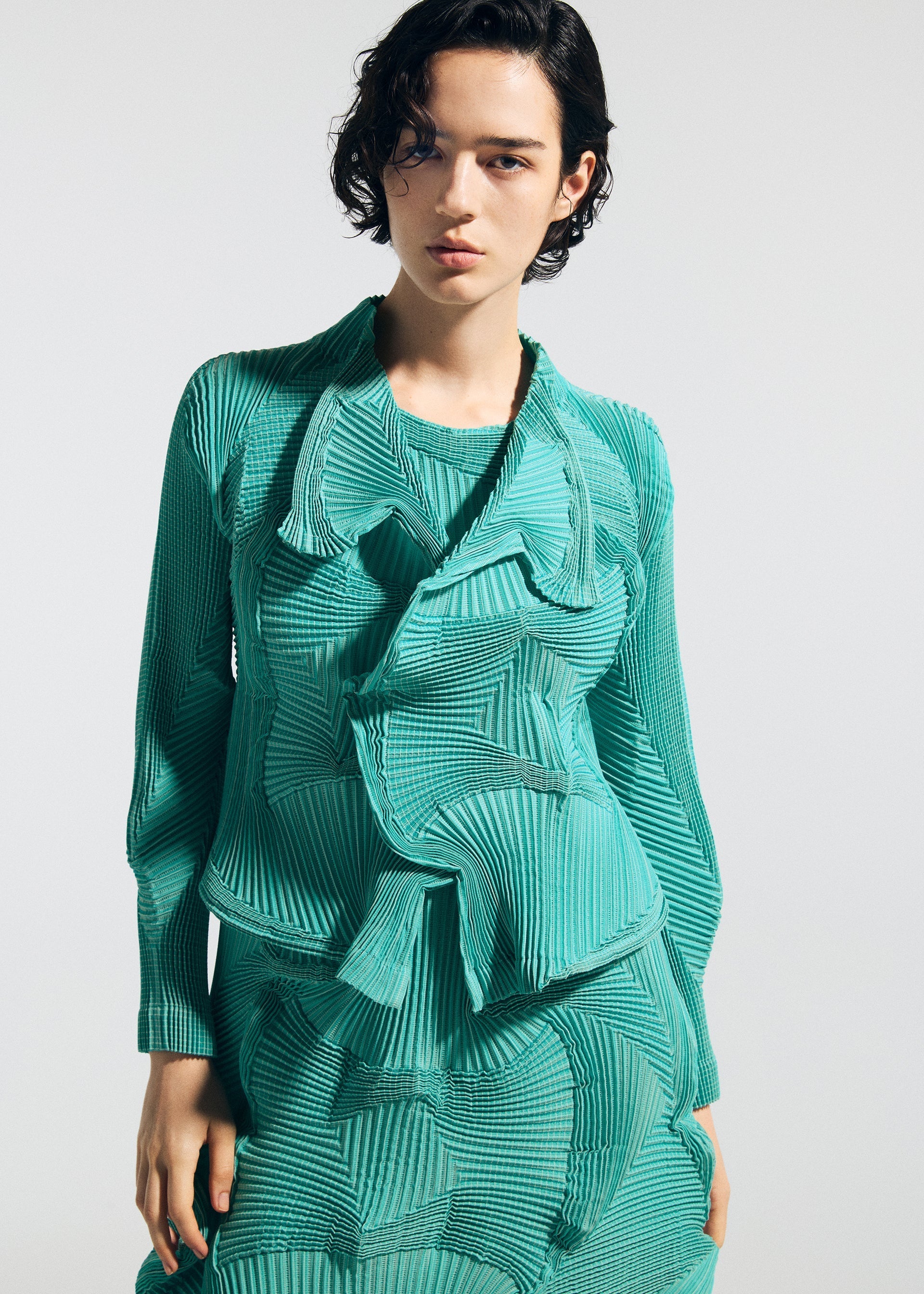 Zoomed view A person with short, dark hair wears the TYPE-O 003 by A-POC ABLE ISSEY MIYAKE—a teal, pleated garment with layered, sculptural details—standing against a plain light background, facing the camera with a neutral expression.