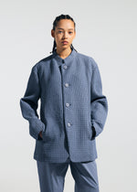 A person with braided hair wears the TYPE-O 014 textured, oversized blue standing collar jacket and matching pants by A-POC ABLE ISSEY MIYAKE, hands in pockets, set against a plain light background.