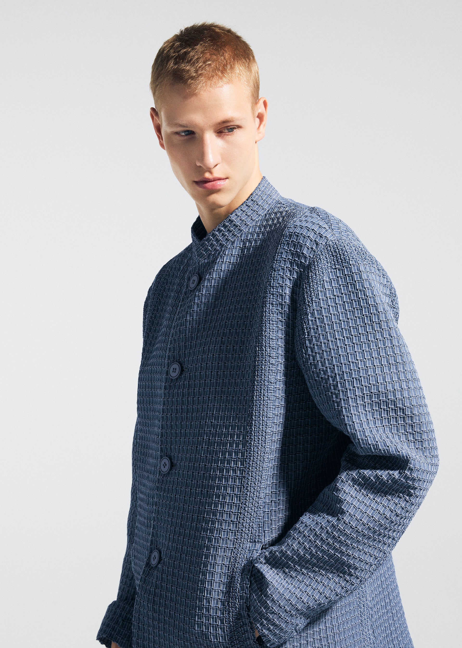 Zoomed view A young person with short blond hair wears the TYPE-O 014 jacket by A-POC ABLE ISSEY MIYAKE, featuring a textured blue standing collar and subtle grid pleats, standing against a plain light background with a neutral gaze.