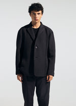 A person with short curly dark hair wears the TYPE-O 014 black tailored collar jacket by A-POC ABLE ISSEY MIYAKE over a black shirt and pants, standing against a light background with a neutral expression.