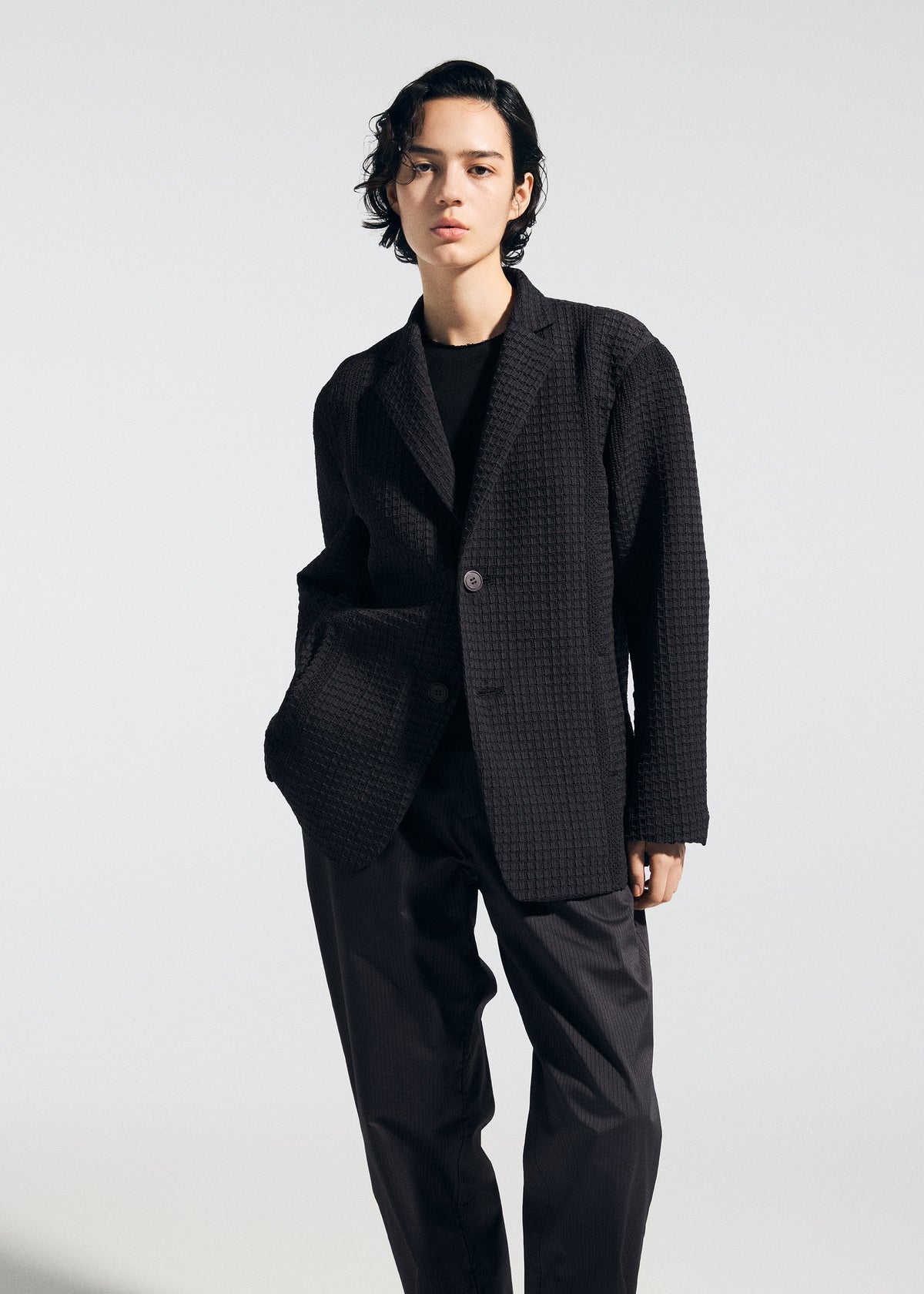 A person with short, wavy dark hair stands against a plain light background, wearing TYPE-O 014 by A-POC ABLE ISSEY MIYAKE: a tailored collar jacket and matching wide-leg pants with grid-shaped pleats, one hand in the pocket, neutral expression. view 6 of 8