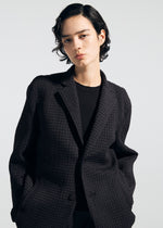 A person with short, wavy dark hair models the TYPE-O 014 black textured tailored collar jacket by A-POC ABLE ISSEY MIYAKE, hands in pockets and facing the camera against a plain light background.