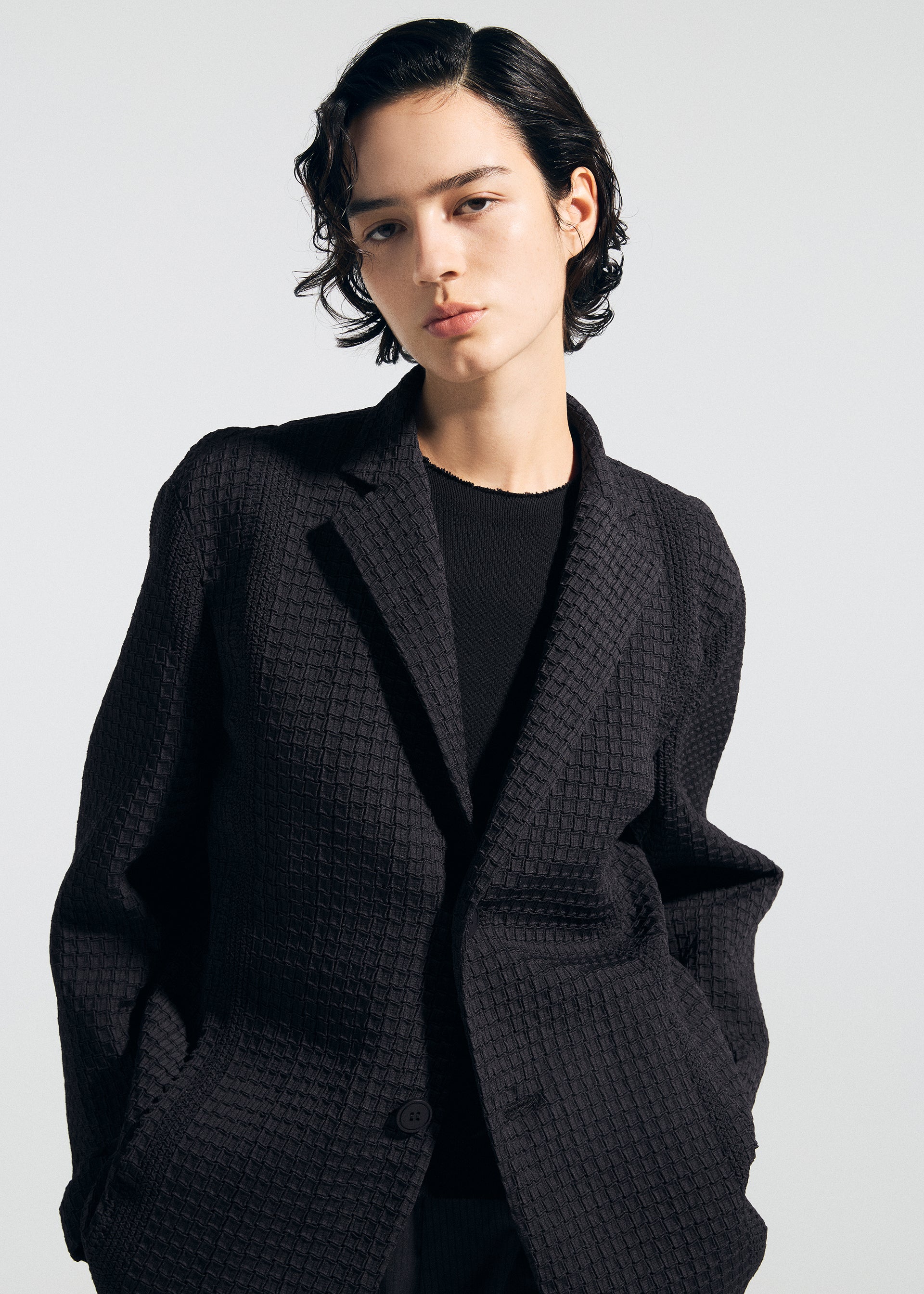 Zoomed view A person with short, wavy dark hair models the TYPE-O 014 black textured tailored collar jacket by A-POC ABLE ISSEY MIYAKE, hands in pockets and facing the camera against a plain light background.