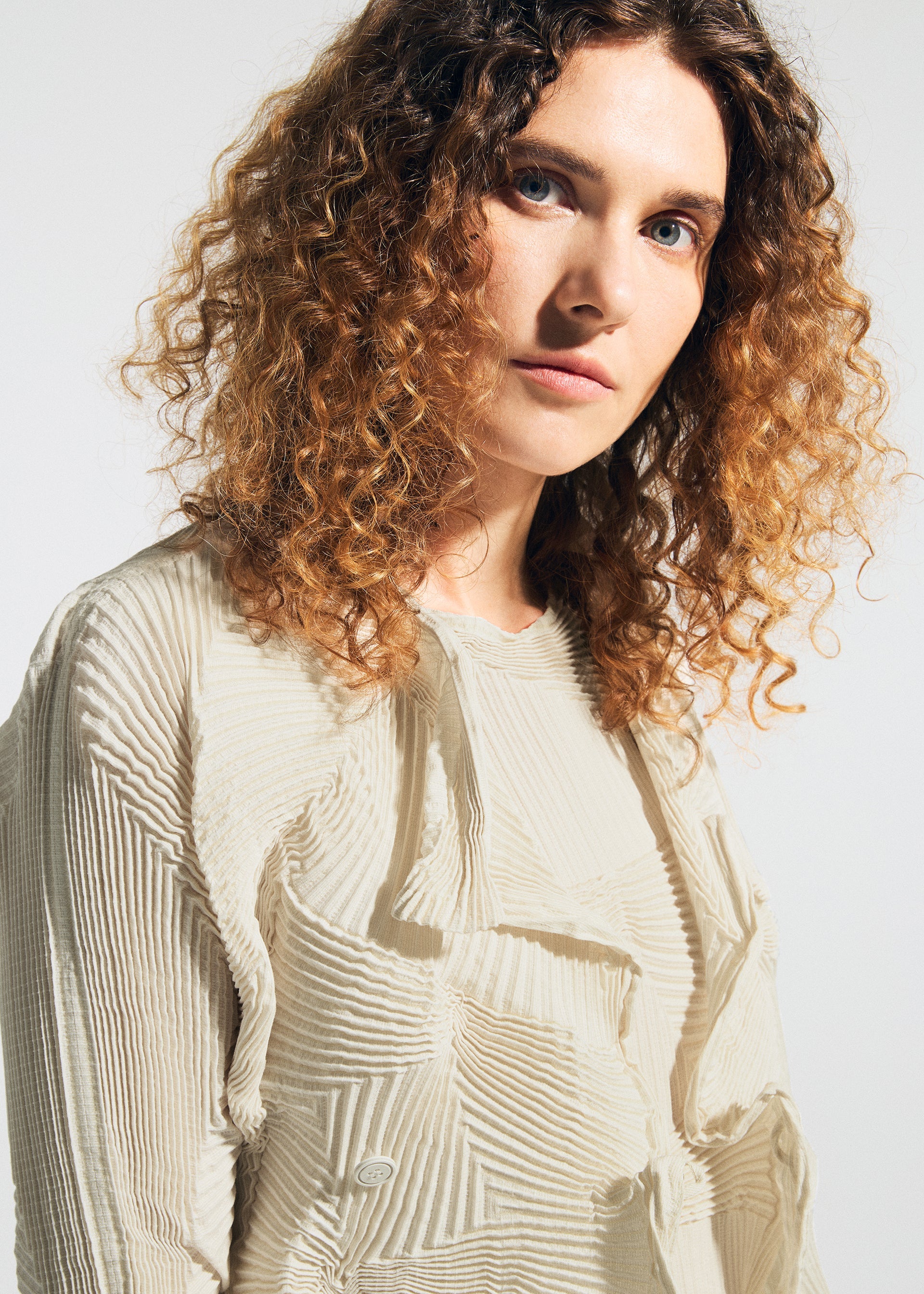 Zoomed view A woman with curly, shoulder-length brown hair and blue eyes wears the TYPE-O 022 jacket by A-POC ABLE ISSEY MIYAKE in cream tones, looking directly at the camera against a plain light background.