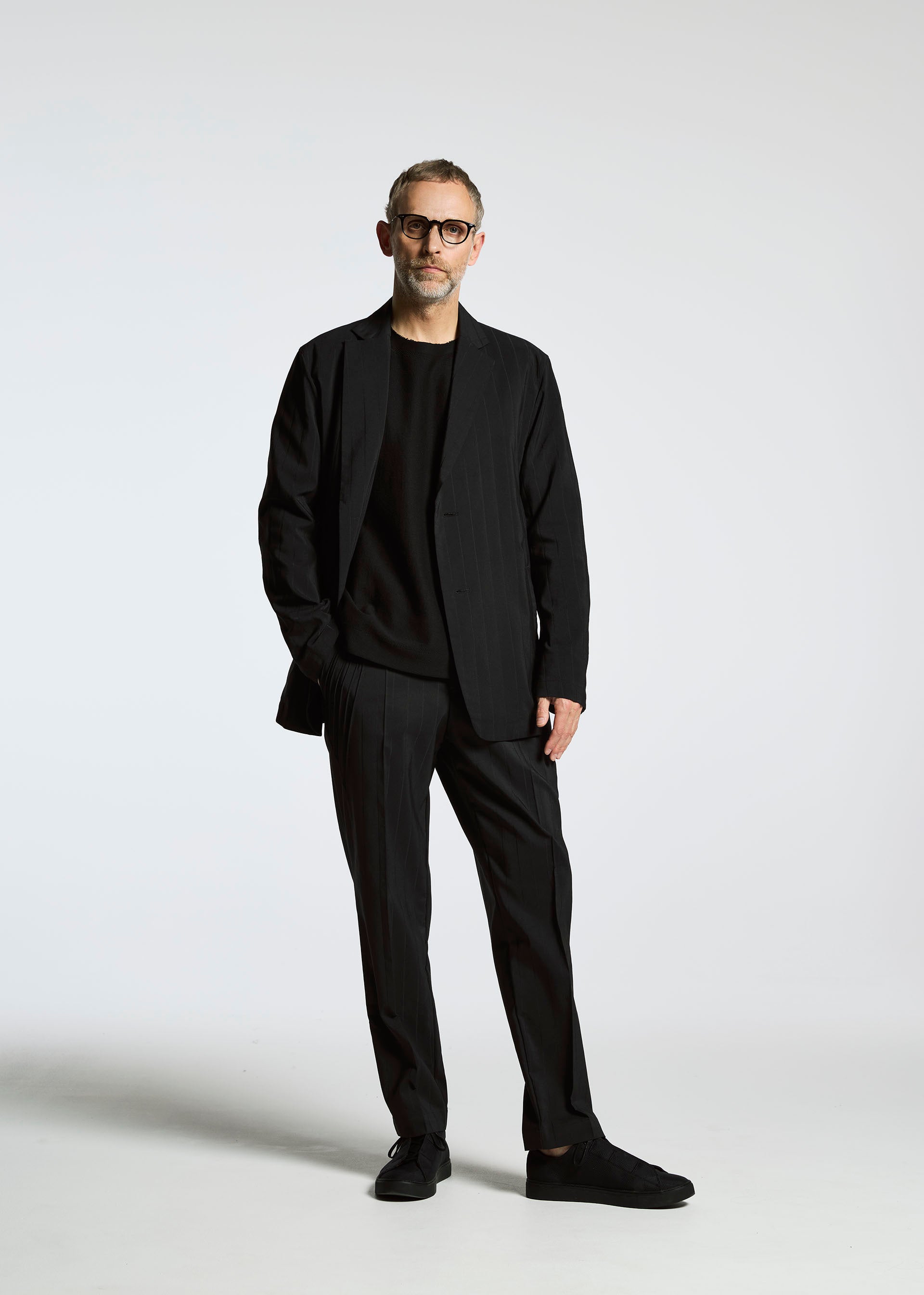 Zoomed view A man with short hair, a beard, and glasses stands against a plain white background wearing the TYPE-Ⅰ 007 jacket by A-POC ABLE ISSEY MIYAKE, paired with a black shirt and black shoes, one hand in his pocket.