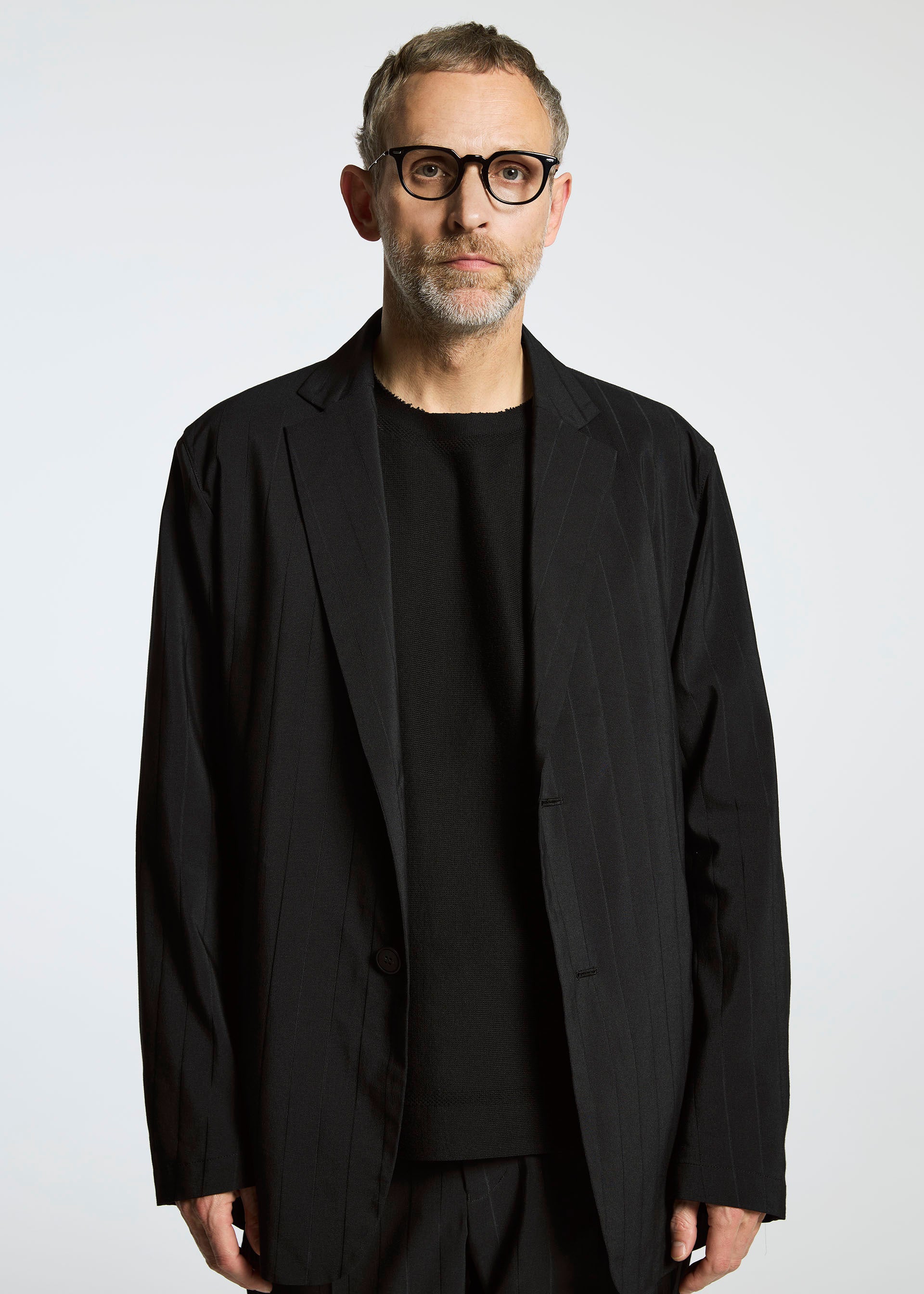 Zoomed view A man with short gray hair, glasses, and a beard wears the TYPE-Ⅰ 007 jacket by A-POC ABLE ISSEY MIYAKE over a black shirt, standing against a plain white background.